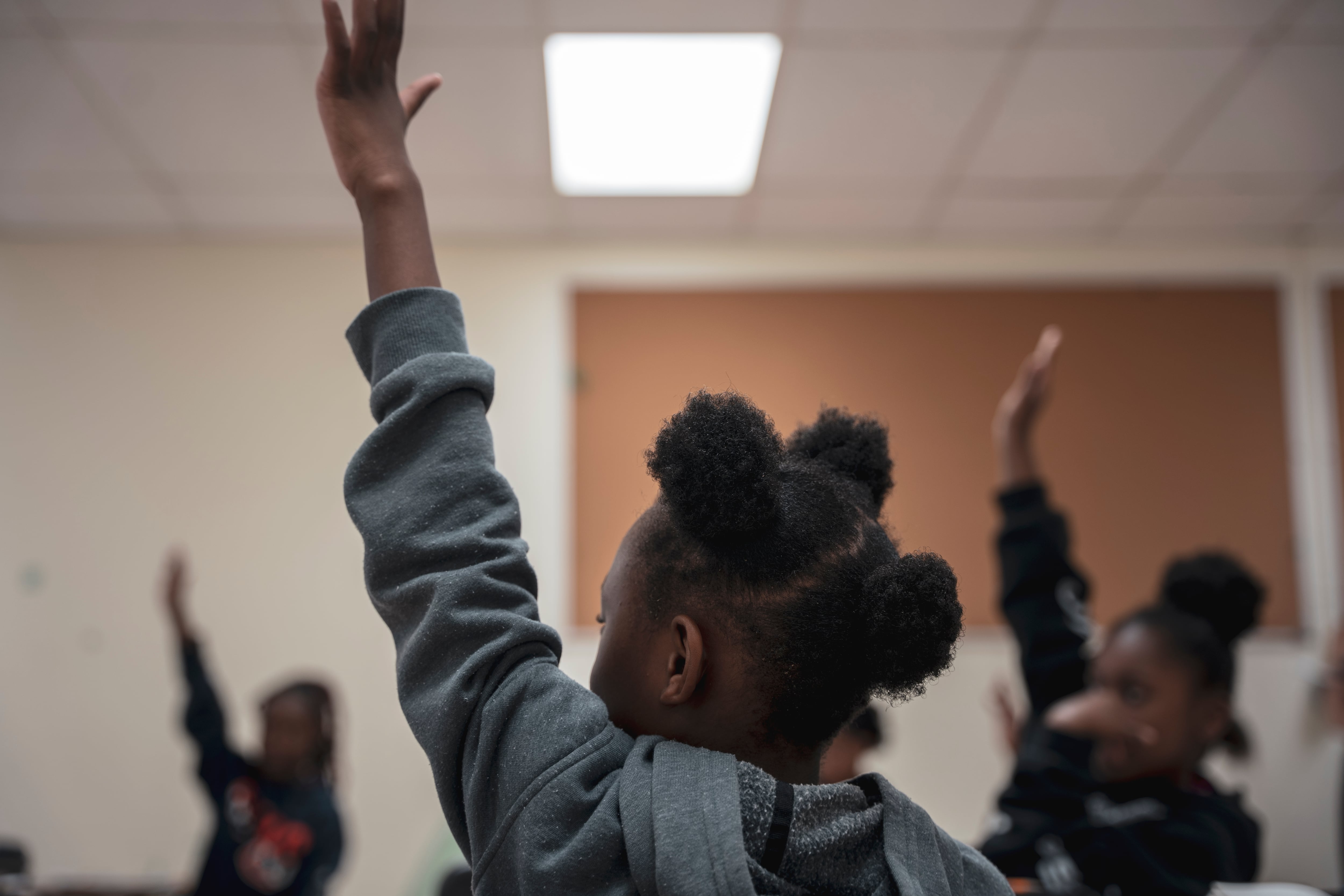 A young girl wearing a grey hoodie raises her hand during class.