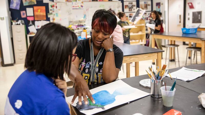 A teacher with a lanyard around her neck works with a student in an art class as they look at a watercolor on a table.
