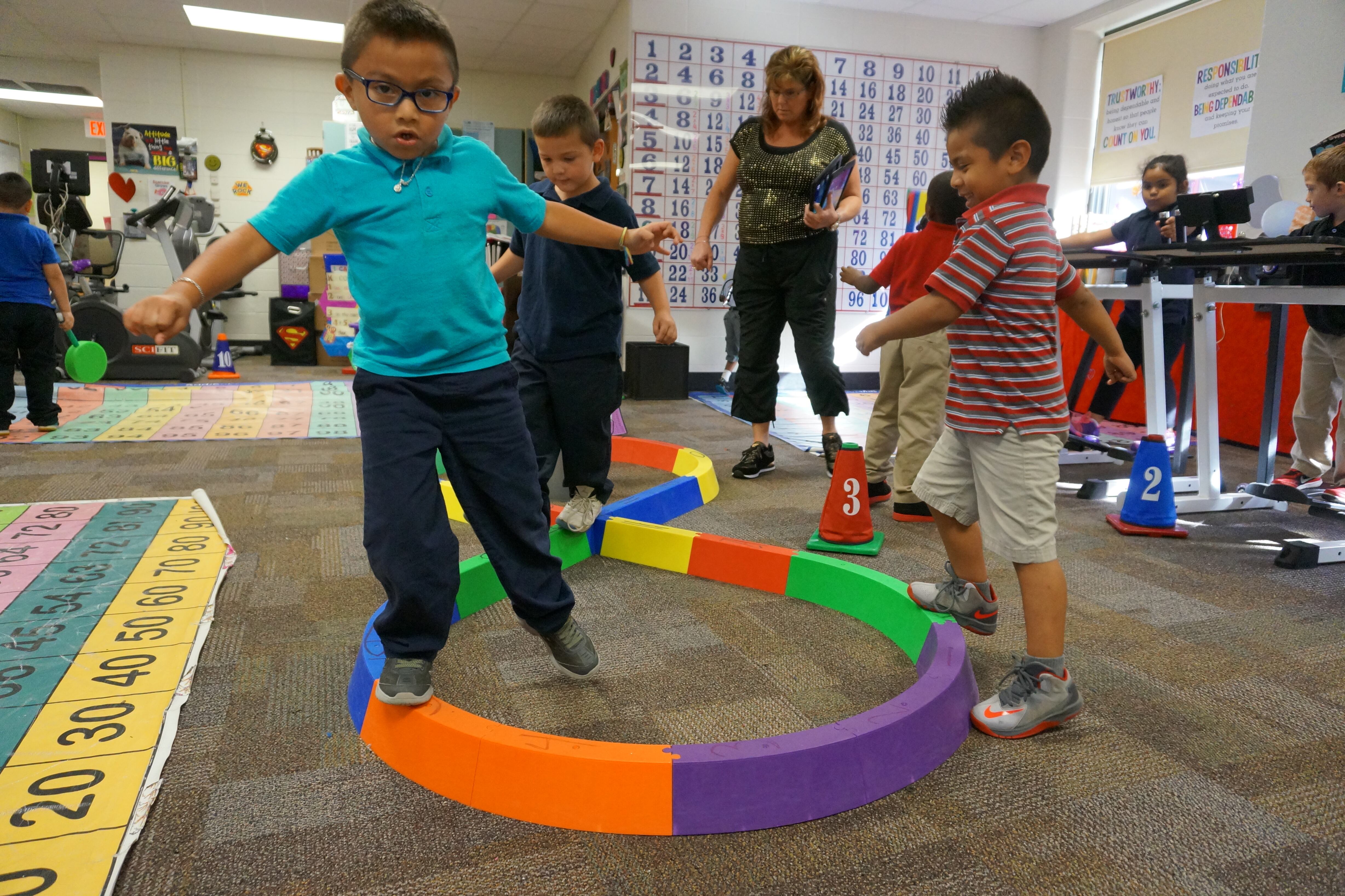 A small child wearing a teal shirt and dark pants and glasses balances on colorful blocks as other students and an adult stand in the background.