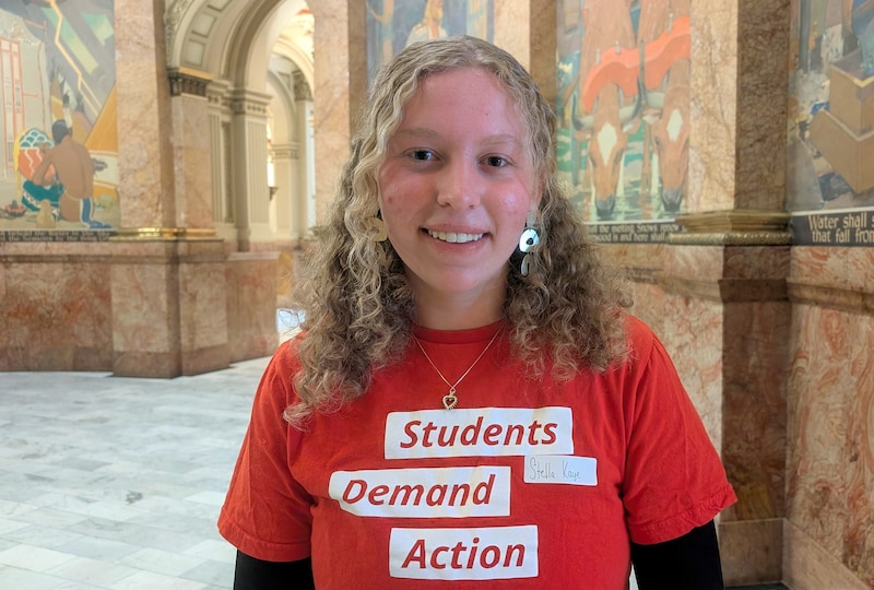 A high school student with long blonde hair and wearing a red shirt standing in the capitol building.