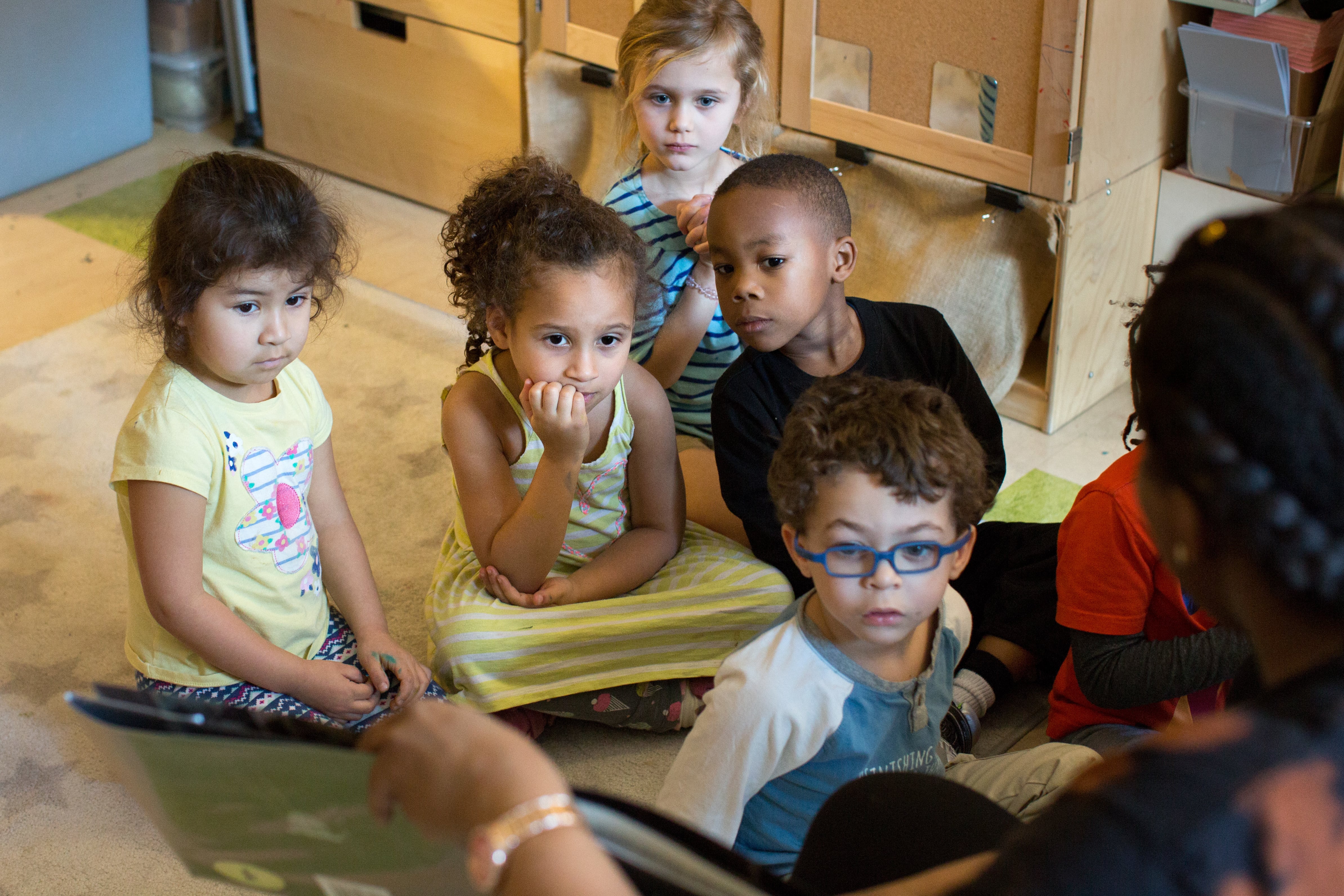 Six little children sit attentively on a rug in a classroom, looking at a book an adult facing them is showing.