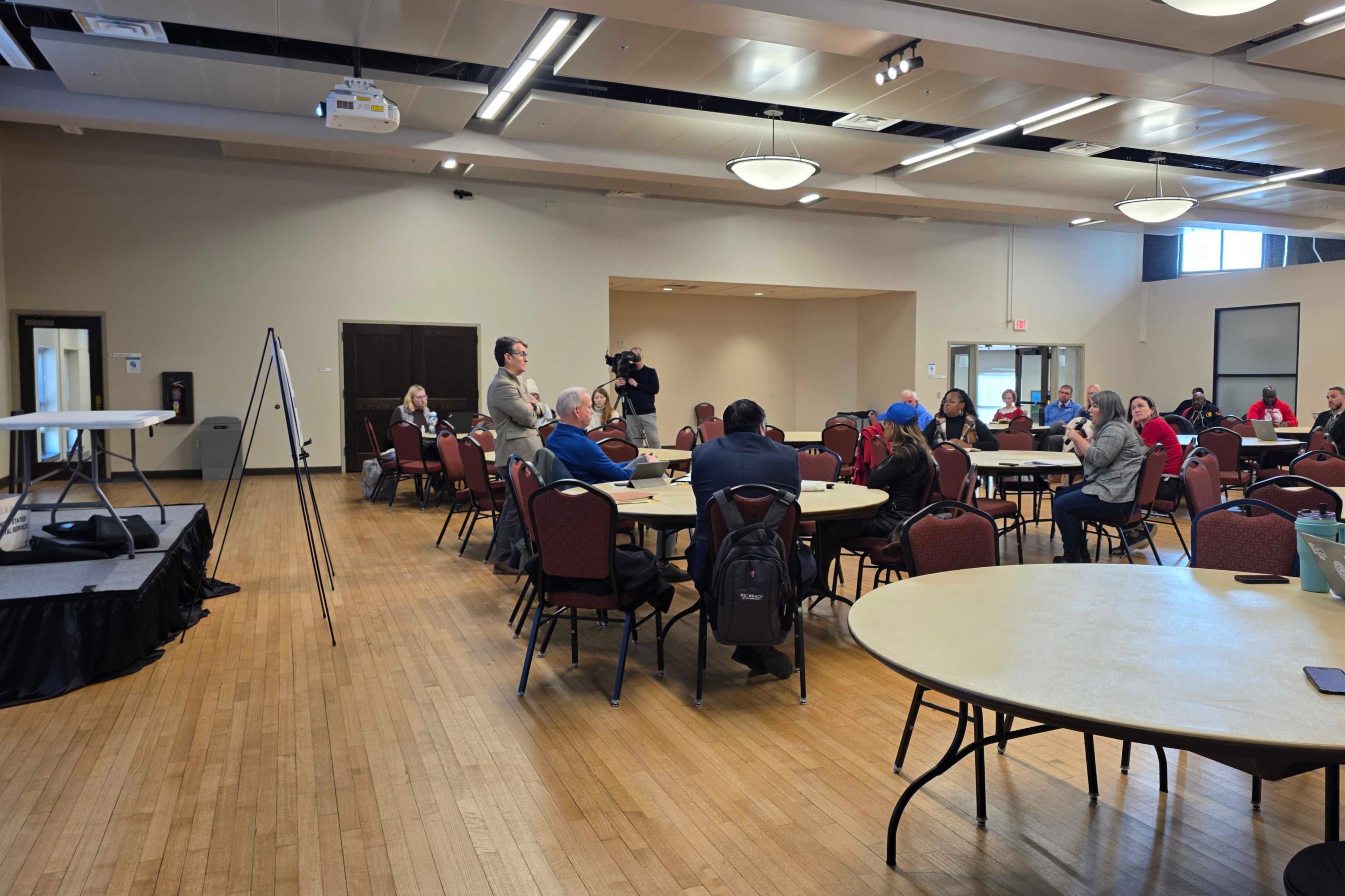 Dozens of people sit at circular tables in a larger room with wooden floors. A projection on the wall shows two diagrams. People watch a woman in a gray sweater talking into a mic while sitting at one of the tables.