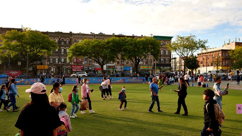 Parents and children, some in face masks, walk across an astroturf field with brick walkup apartment buildings in the background.