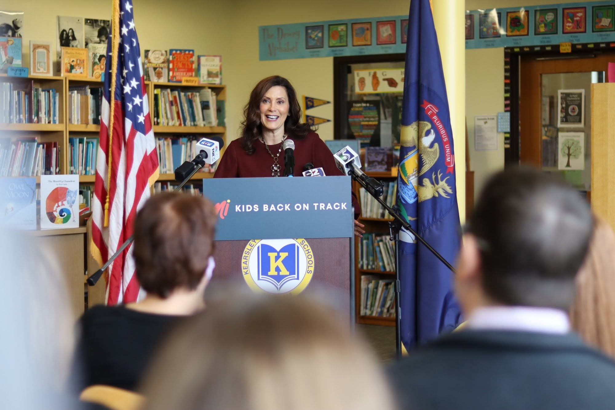 Gov. Gretchen Whitmer speaks into a microphone at a podium with the words MI kids back on track in front of a united states flag as a crowd looks on.