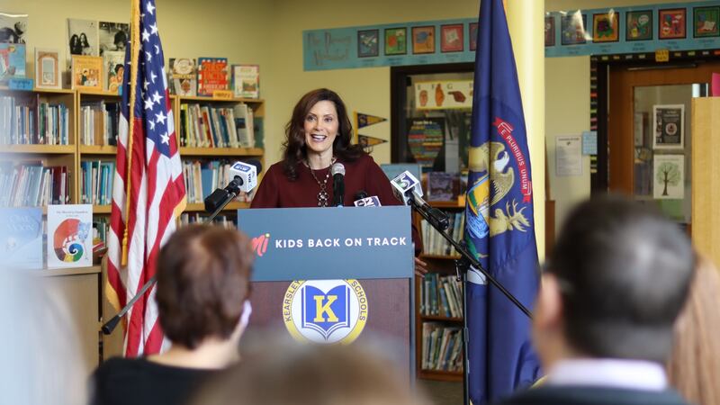 Gov. Gretchen Whitmer speaks into a microphone at a podium with the words MI kids back on track in front of a united states flag as a crowd looks on.