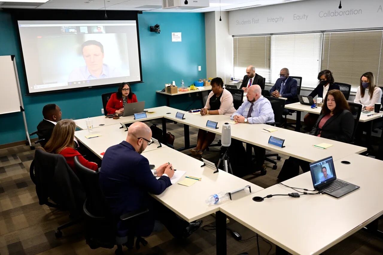 A group of people sit around tables that form a U. A screen at the front of the room shows a person’s face.