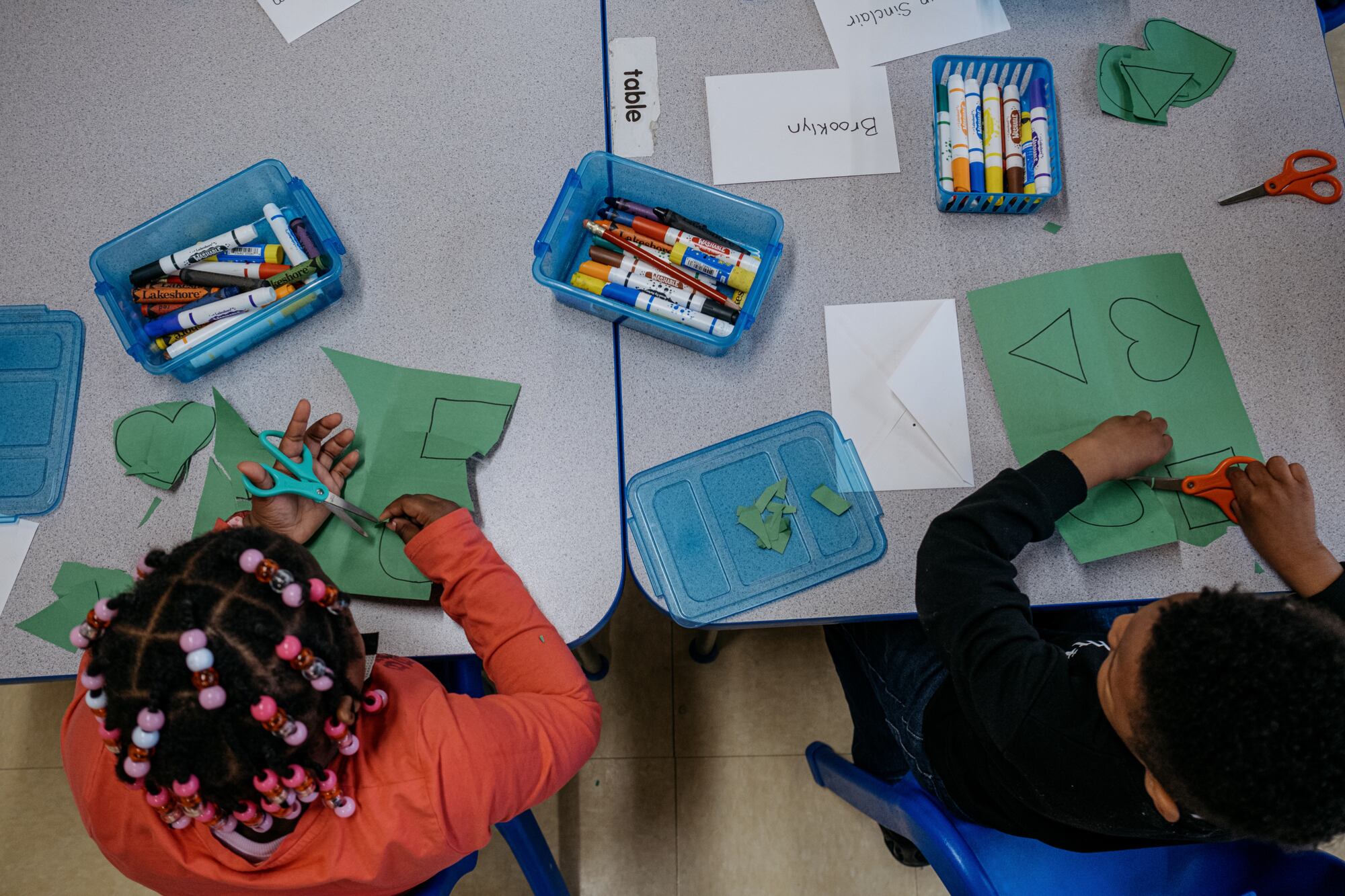 A bird's eye view of two young students sitting at a table cutting out shapes from construction paper.