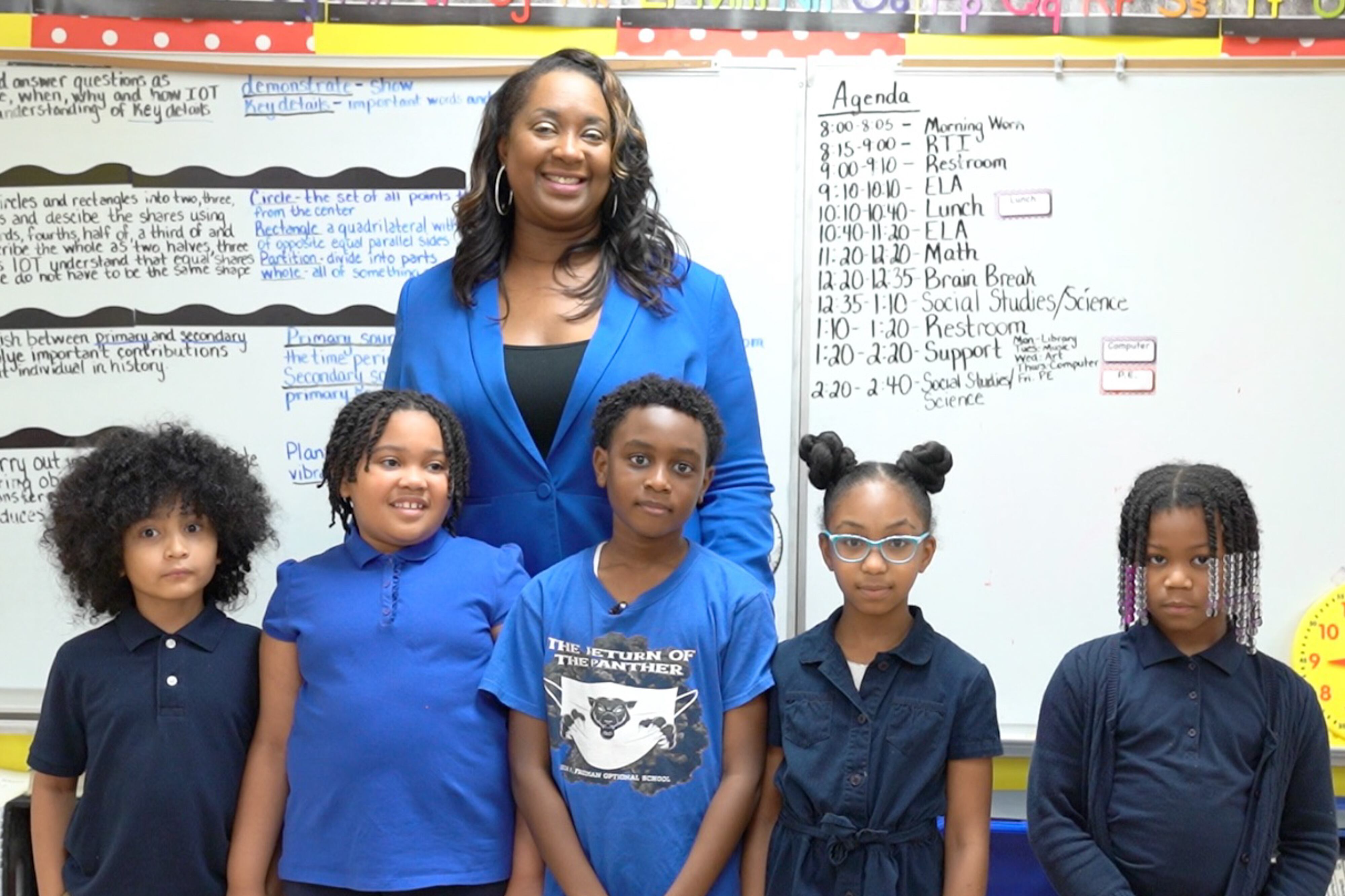 A teacher stands behind five children in a classroom against the backdrop of a whiteboard.