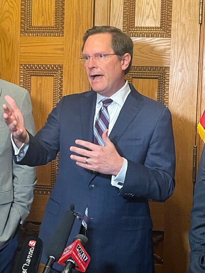 A man wearing a dark suit and glasses speaks to a crowd while standing in front of a wooden door.