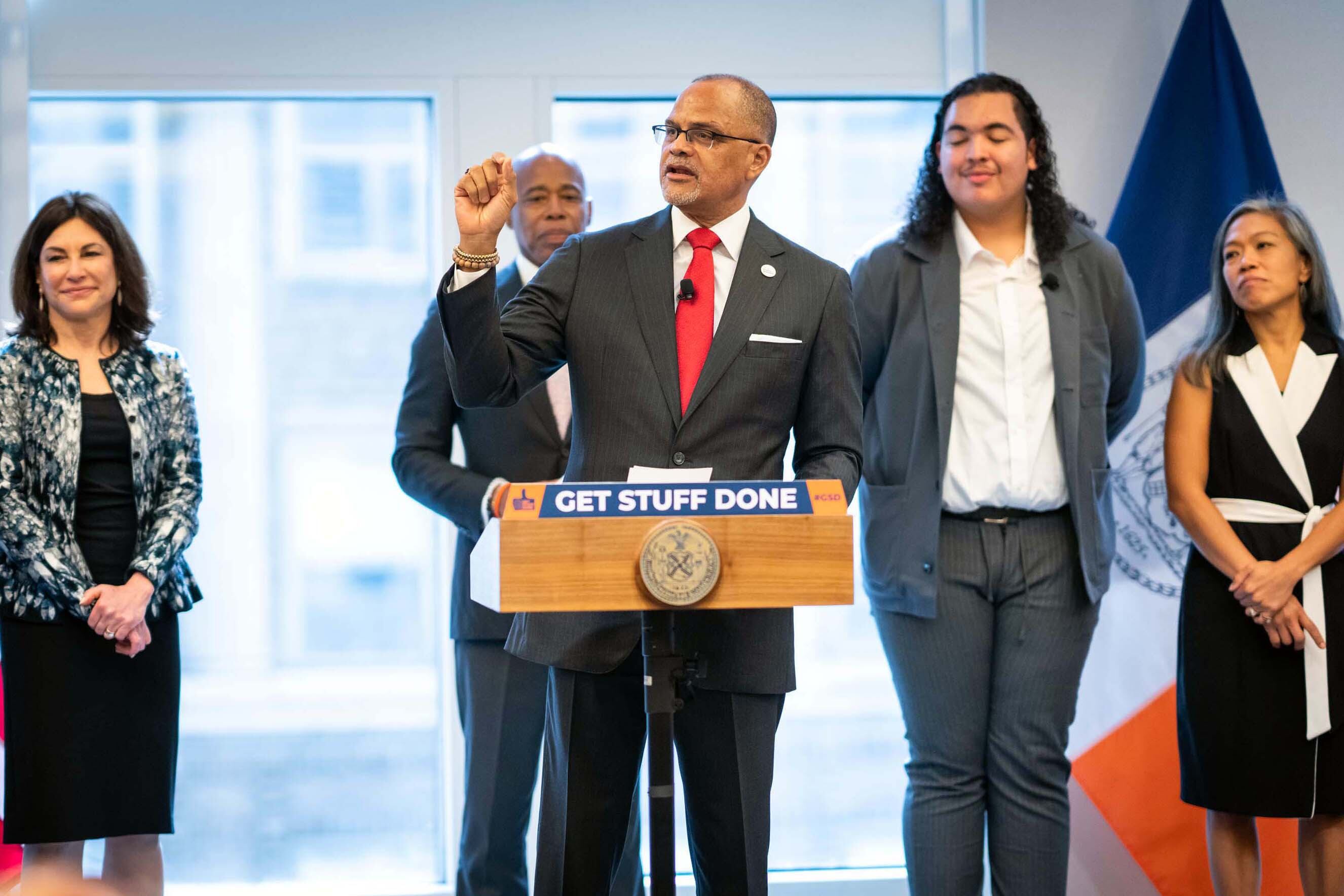 A man in a suit and red tie stands at a wood podium surrounded by onlookers.