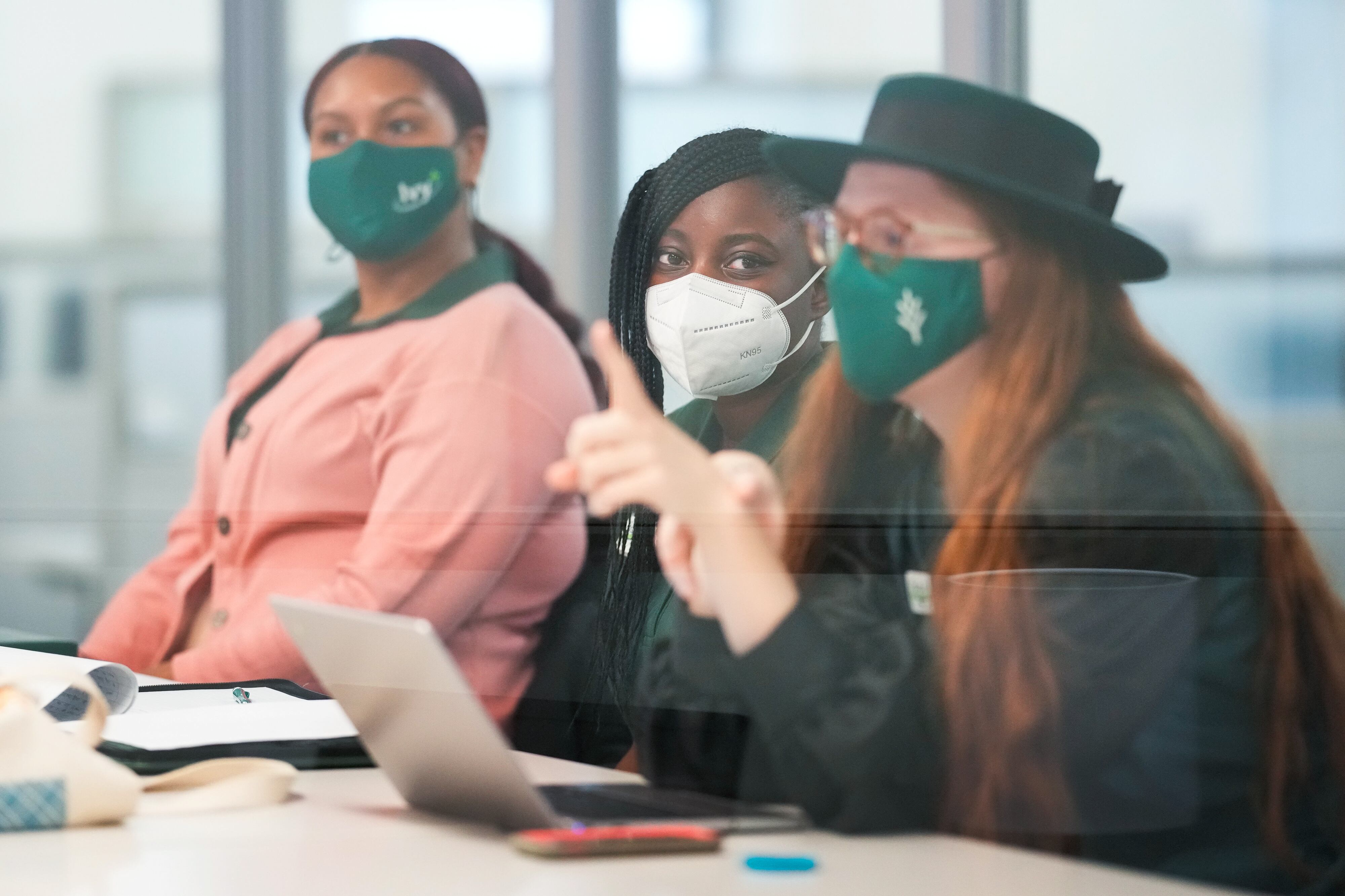 Three college students, wearing protective masks, participate in a meeting at school. There is a slight reflection off of a pane of glass in the foreground.