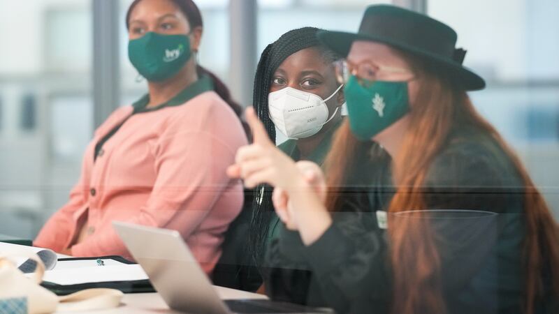 Three college students, wearing protective masks, participate in a meeting at school. There is a slight reflection off of a pane of glass in the foreground.