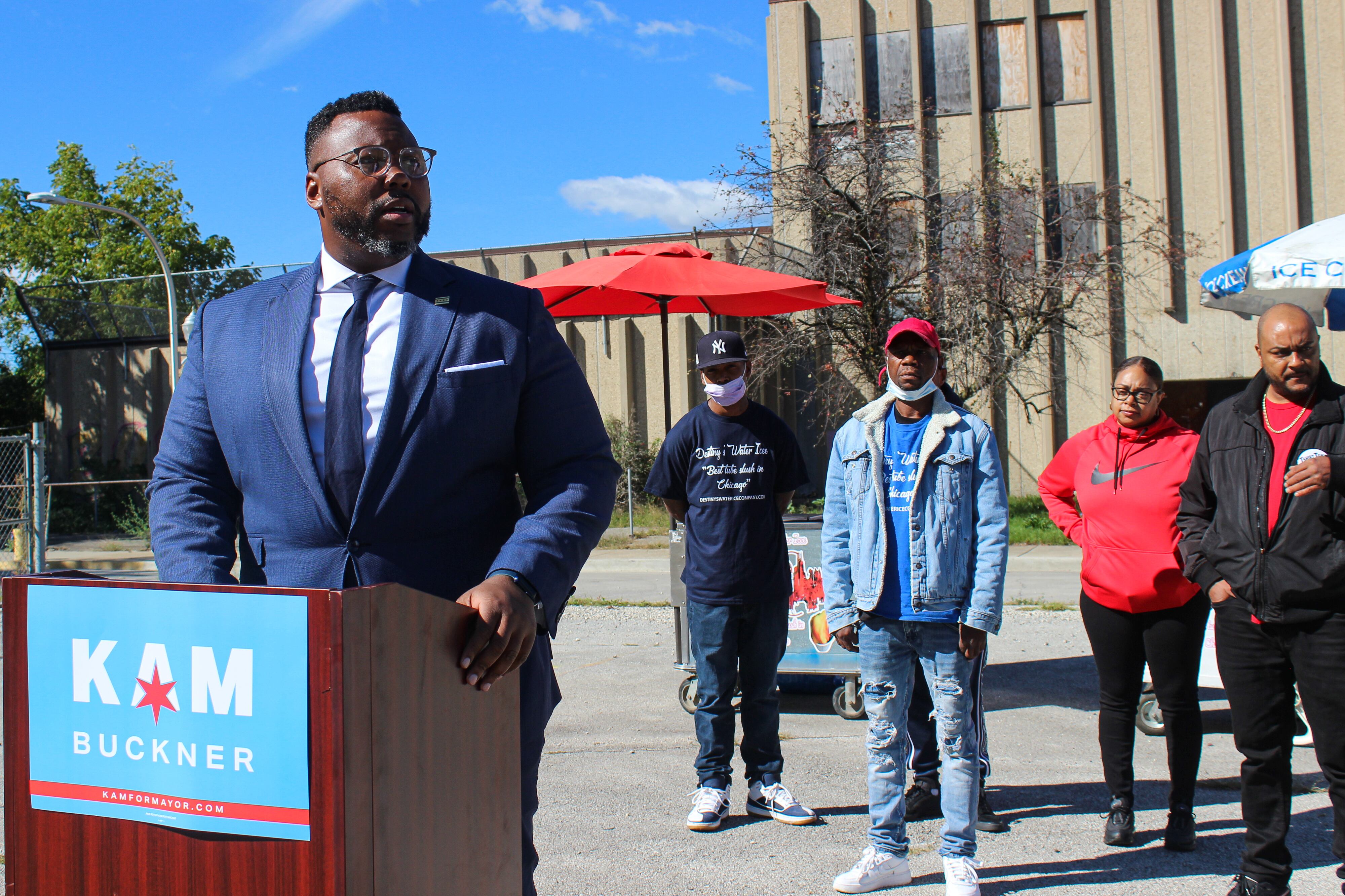A man stands at a podium alongside a handful of supporters in a vacant lot across from an abandoned elementary school.