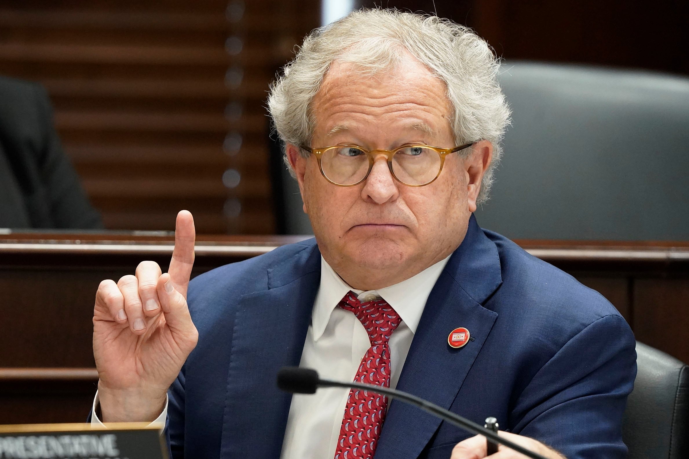 A white man with short, white hair and wearing a suit holds one finger up while sitting at a wooden podium.