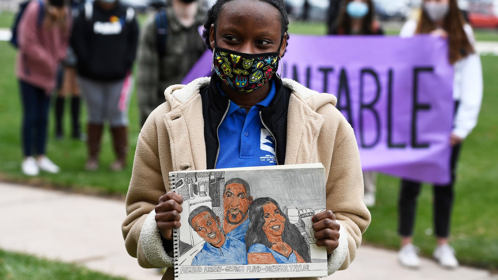 A young woman wearing a cream colored jacket, blue shirt and cloth mask holds a drawing of Ahmaud Arbery, George Floyd and Breonna Taylor. Students hold a purple banner behind her.