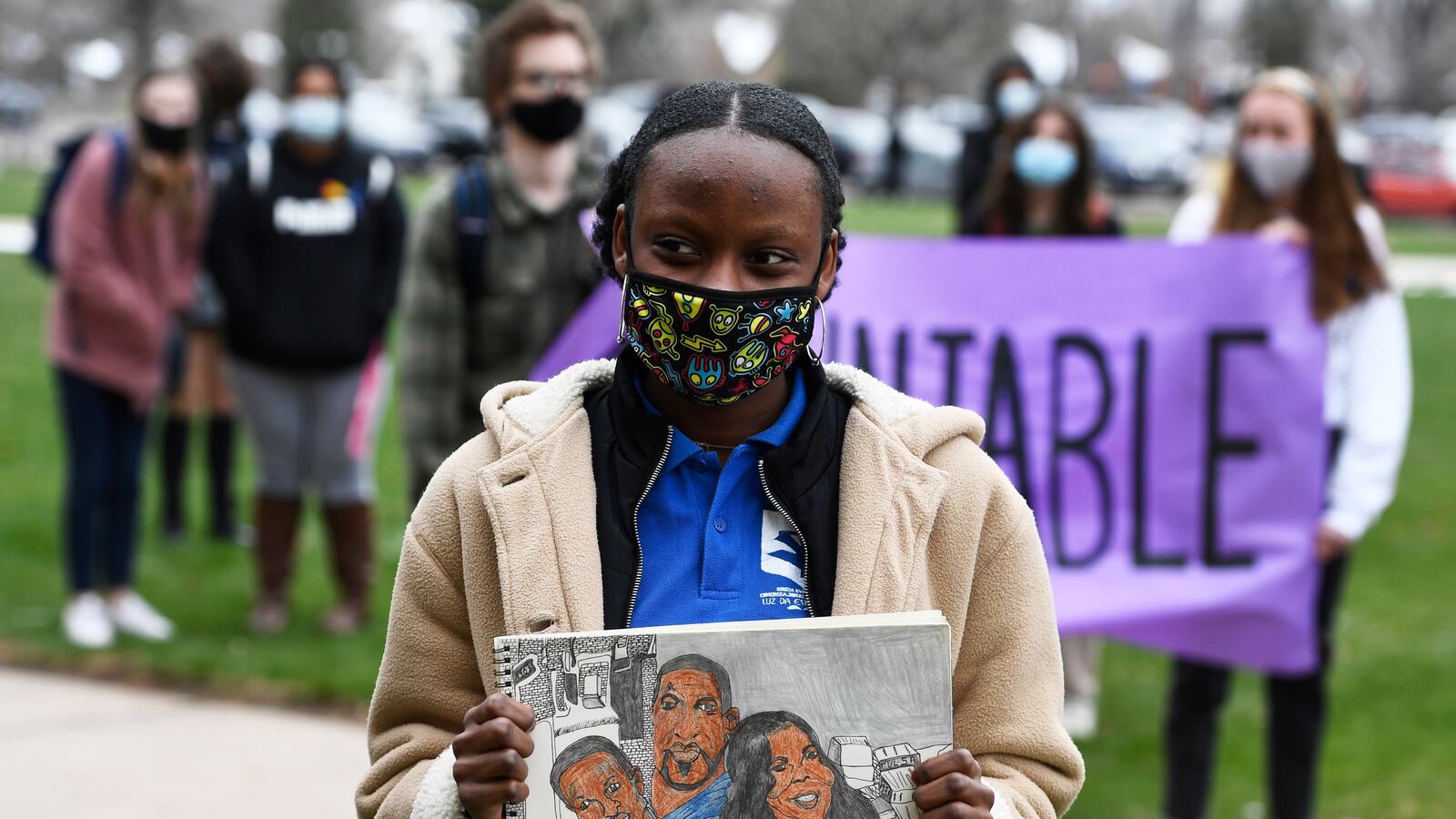 A young woman wearing a cream colored jacket, blue shirt and cloth mask holds a drawing of Ahmaud Arbery, George Floyd and Breonna Taylor. Students hold a purple banner behind her.