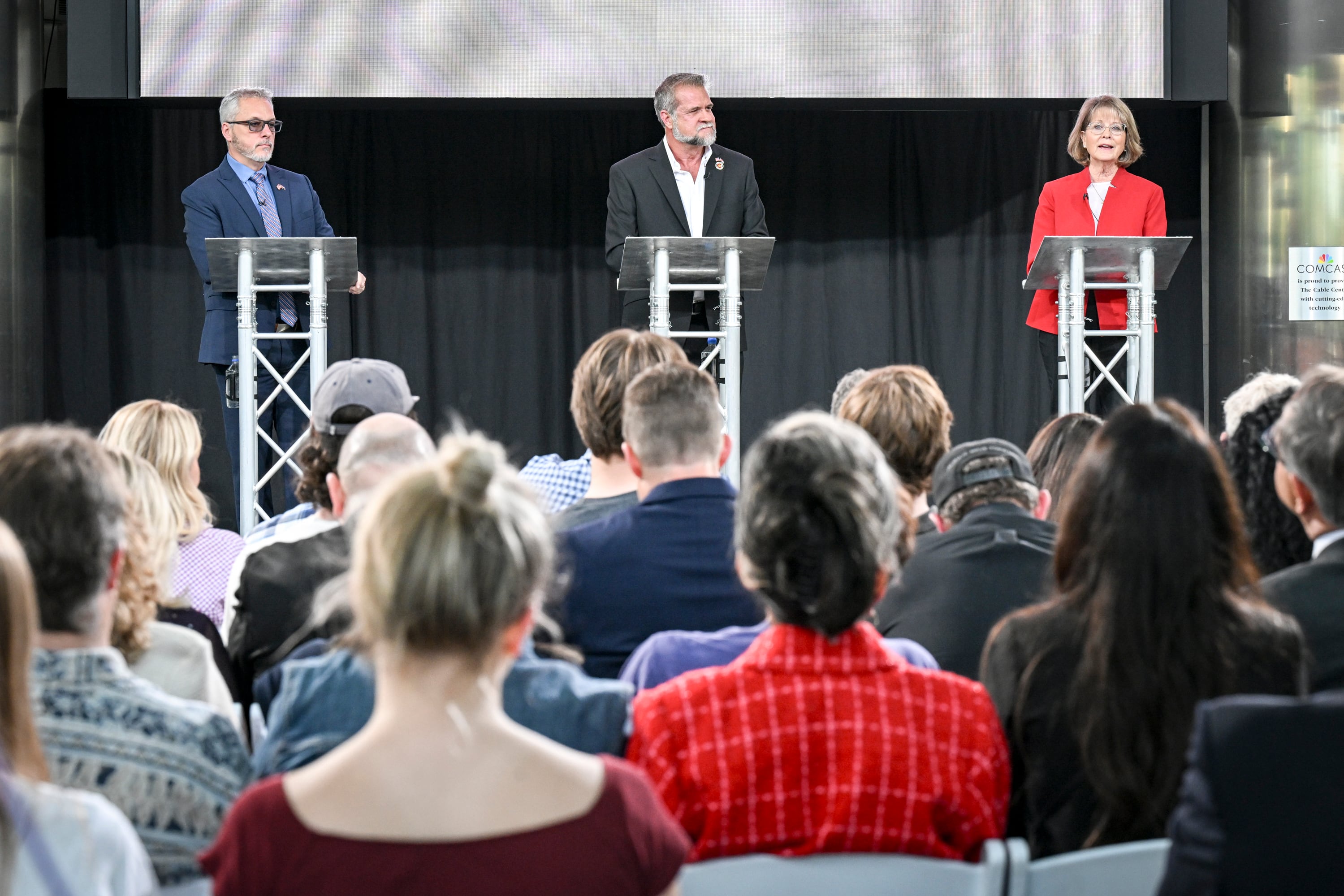 A photograph of two white men and a white woman in suits speaking from behind podiums on a stage with a group of audience sitting in the foreground.