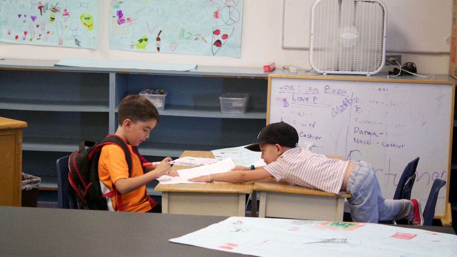 Two elementary students sit at a pair of desks in a classroom. One student is reaching over his desk toward the other student.