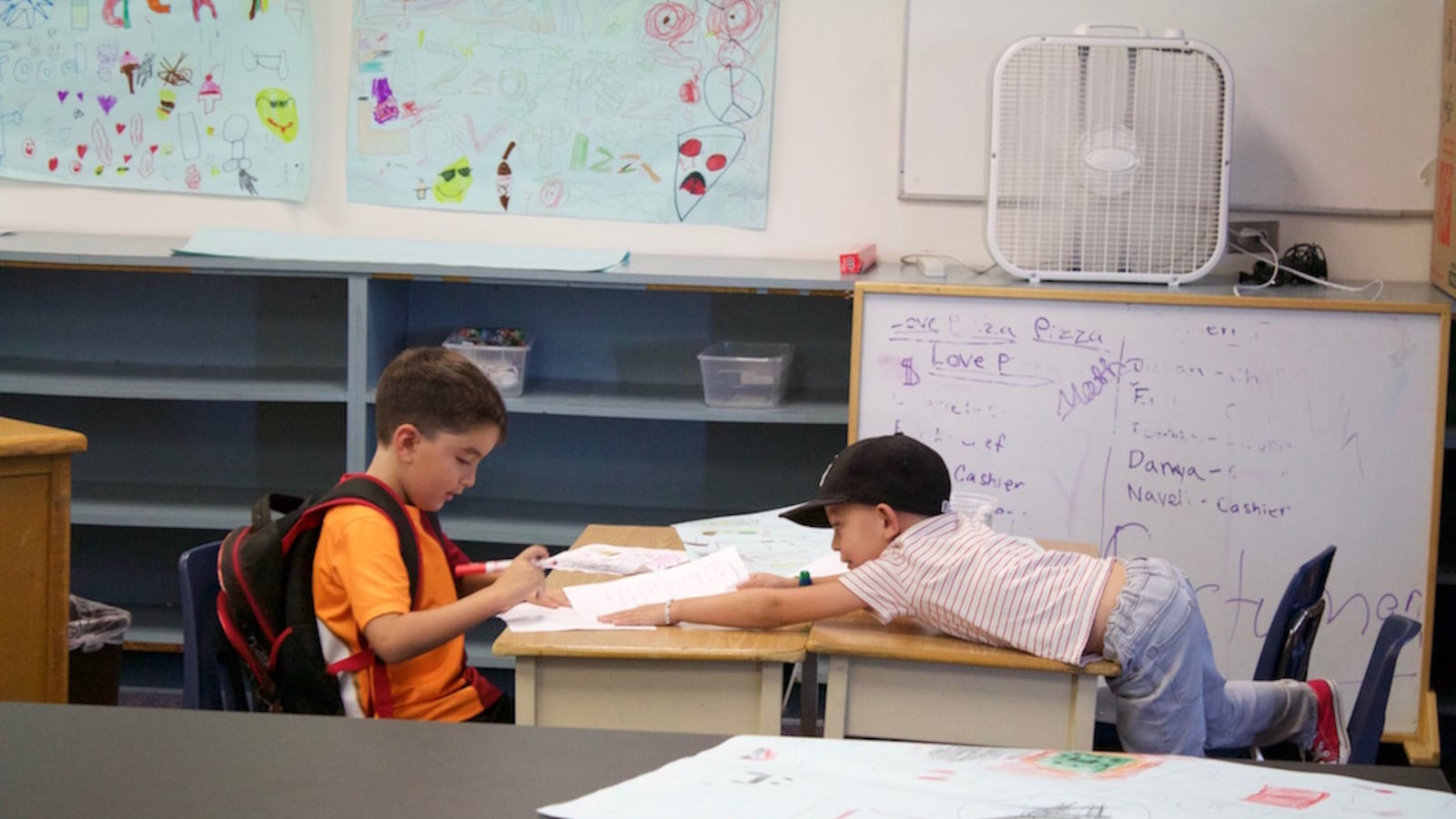 Two elementary students sit at a pair of desks in a classroom. One student is reaching over his desk toward the other student.