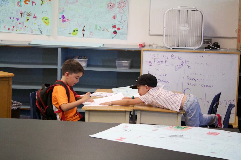 Two elementary students sit at a pair of desks in a classroom. One student is reaching over his desk toward the other student.