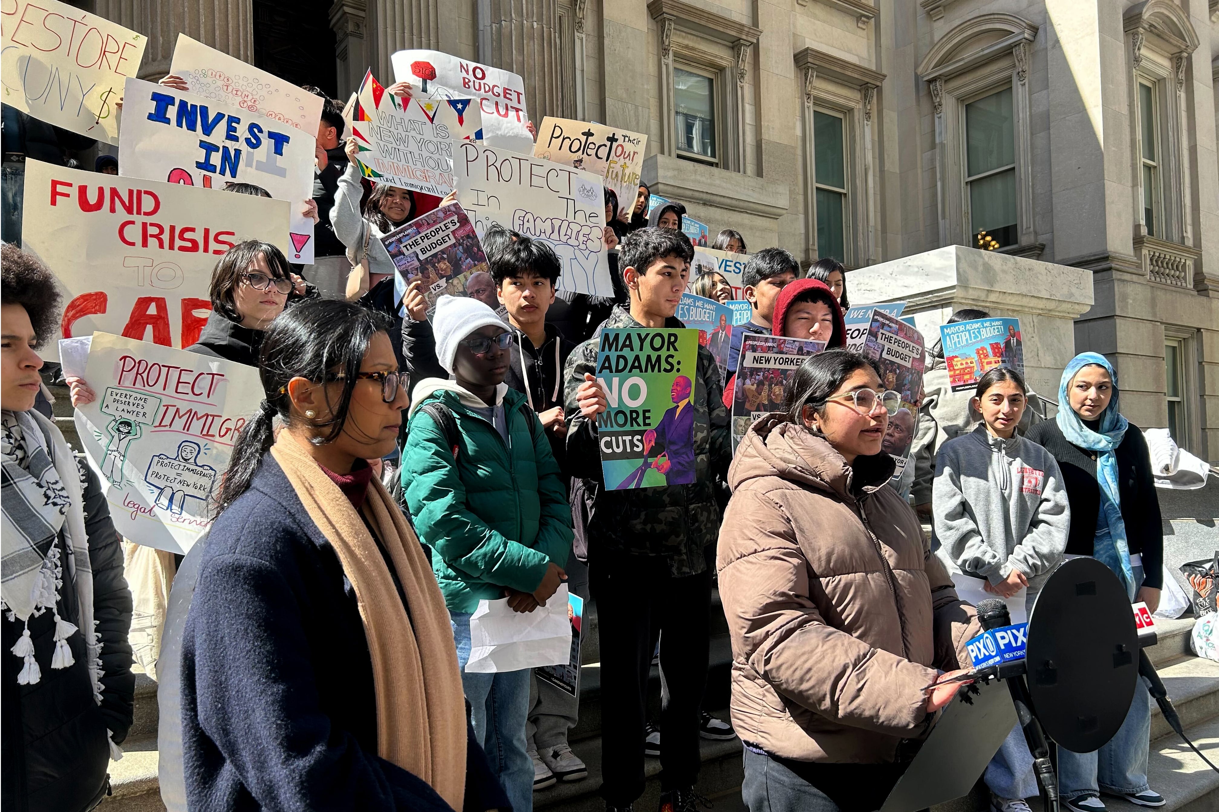 A large group of high school students stand together in a group on stone steps outside of a large stone building, some are holding signs.