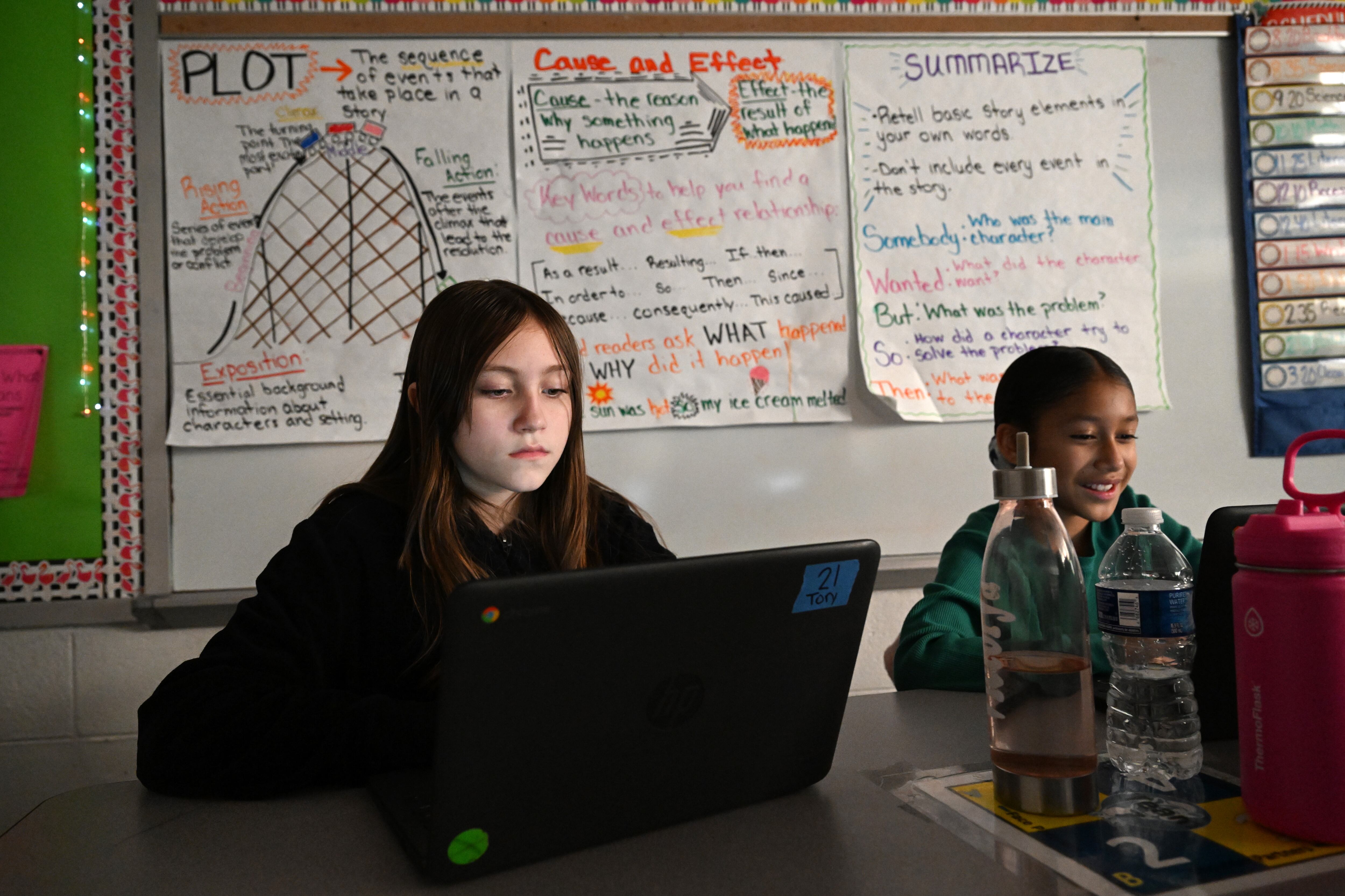 Students sitting at desks look at their laptops. Their faces are slightly illuminated by their screens. Educational posters are on the wall behind them.