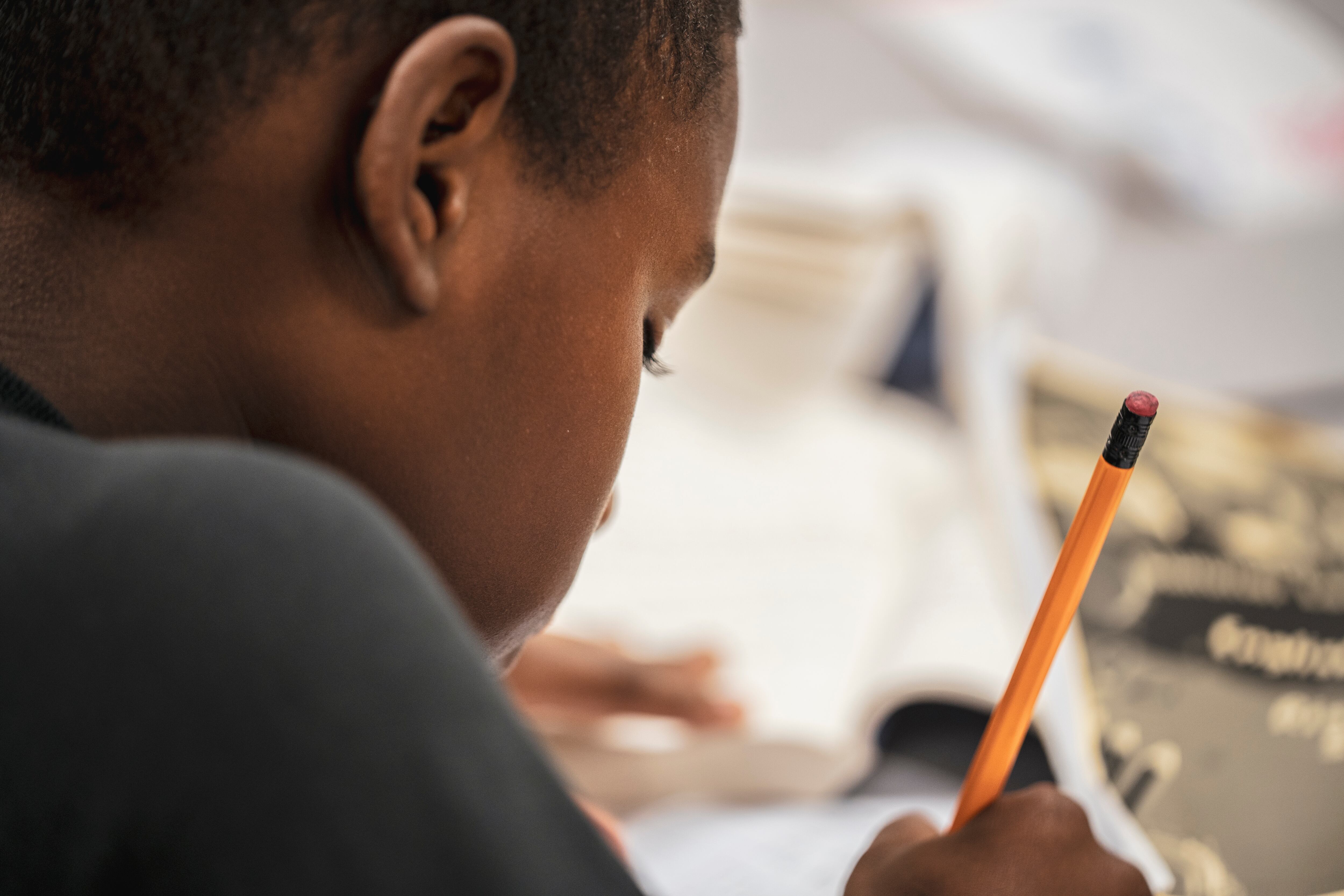 A close up of a young Black boy holding a pencil and writing on a piece of paper while sitting at a desk.