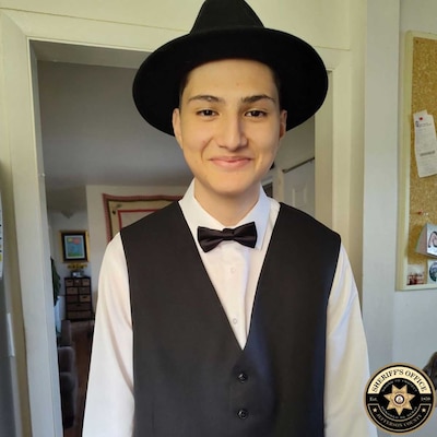 A photograph of a teenage boy wearing a black hat an a suit vest and bowtie posing for a photograph.