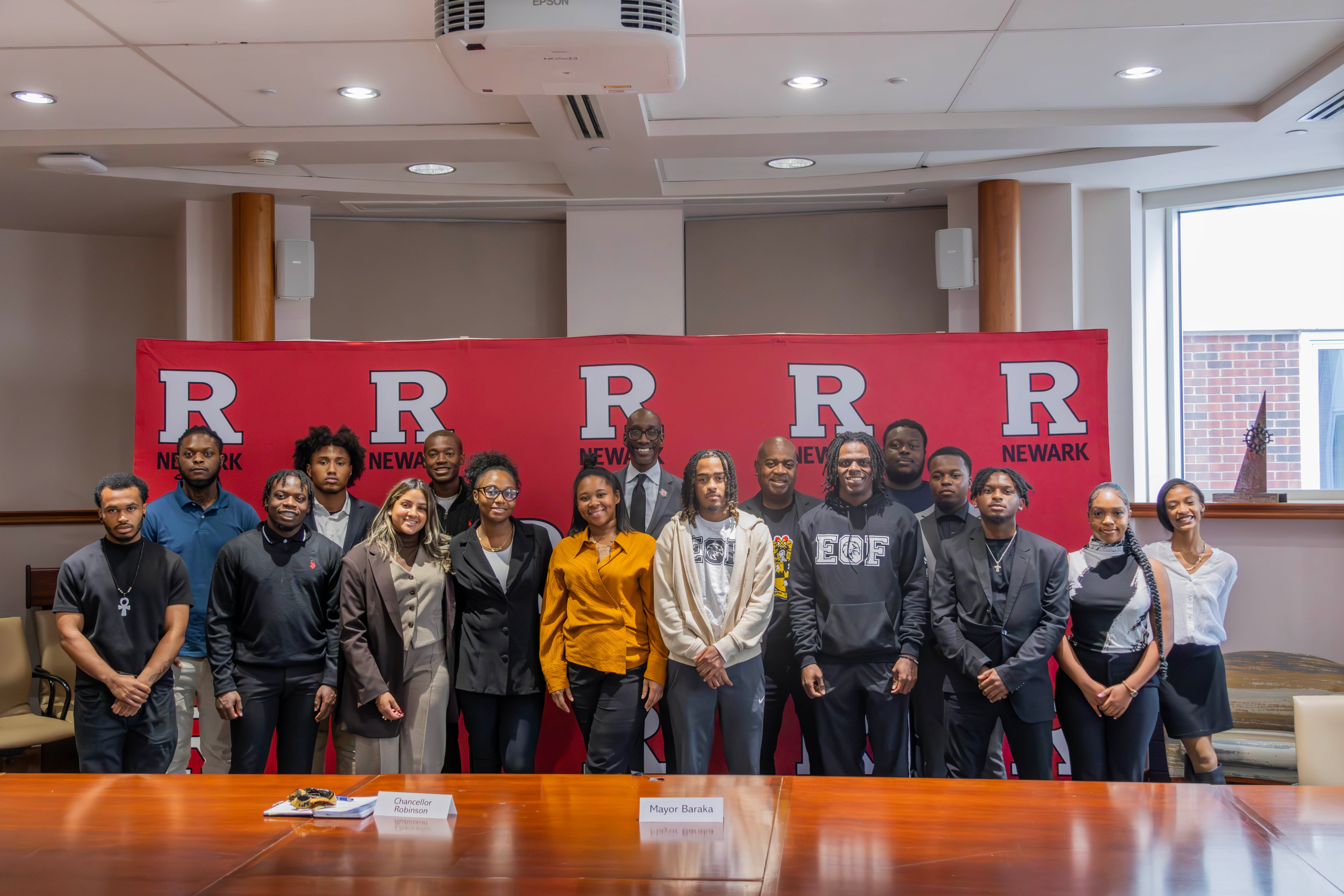 A group of university students dressed in casual business attire stand with city and university officials in front a step-and-repeat backdrop with the Rutgers University logo.