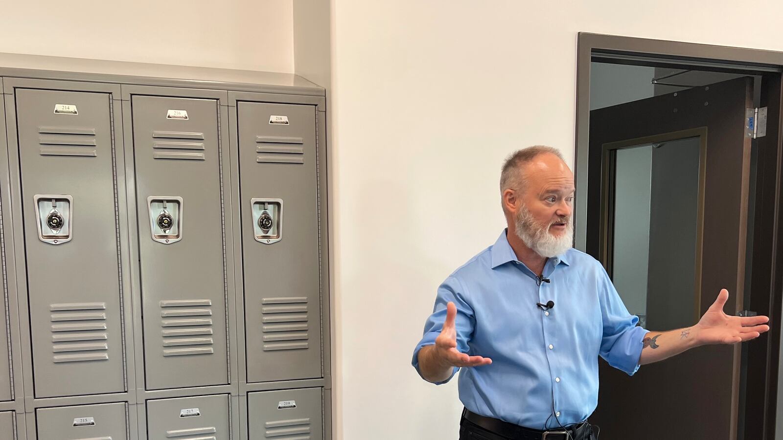 A man in a blue shirt holds out his hands while explaining the building with lockers to the left of him.