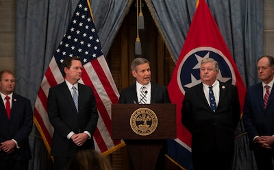 A man wearing a business suit speaks into a microphone behind a podium, surrounded by four other men wearing business suits.