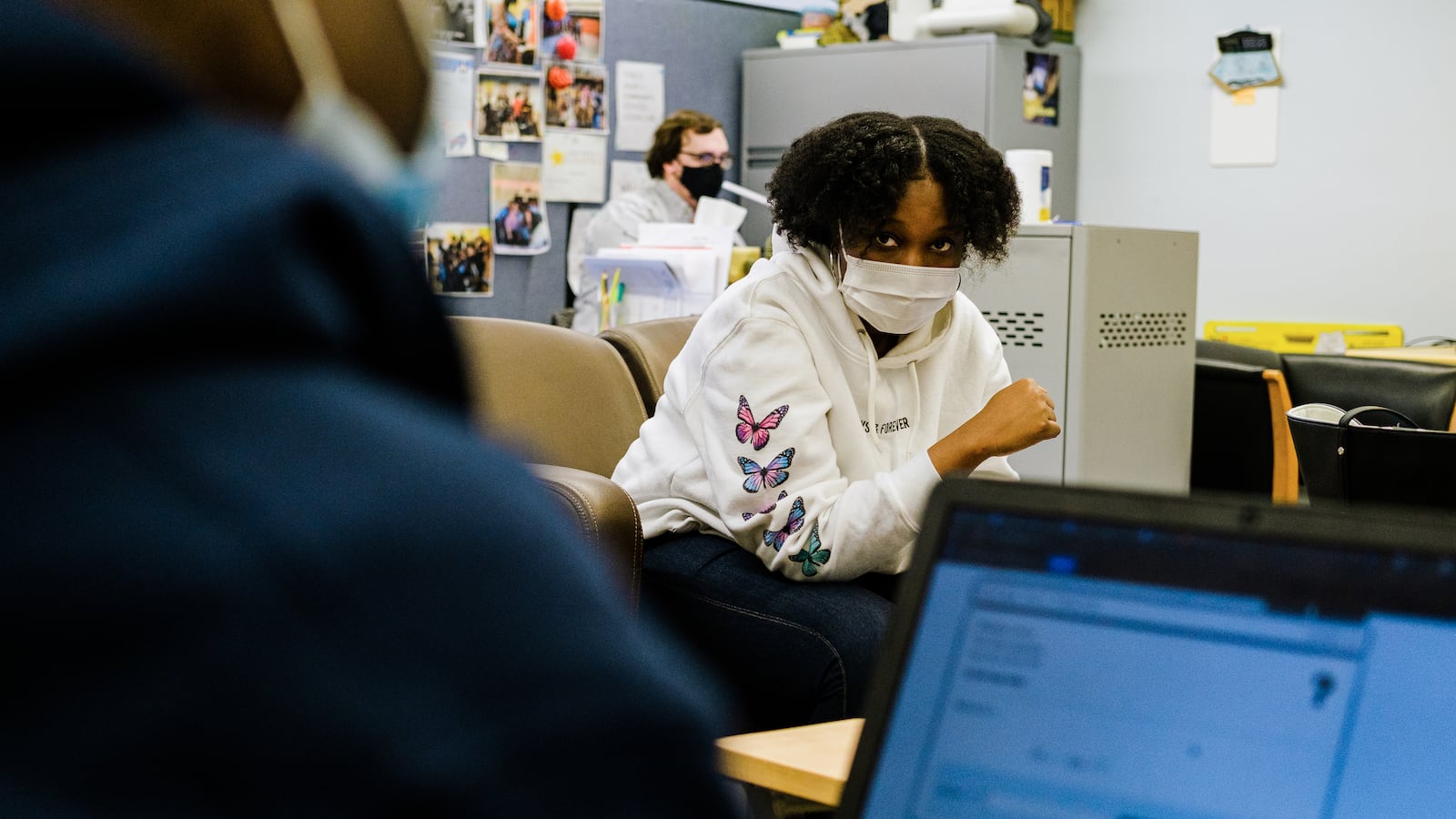 A young woman wearing a protective mask, white hoodie with blue and pink butterflies sits across from a young man wearing a blue hoodie working on a laptop. There is a male teacher sitting behind a desk in the background.