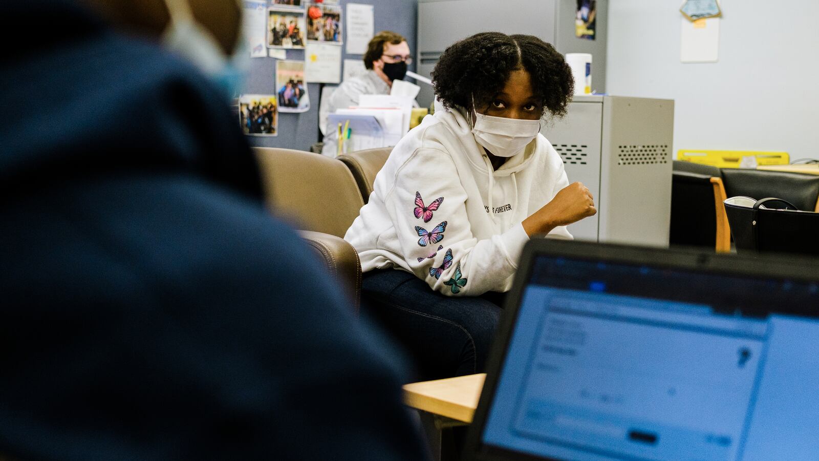 A young woman wearing a protective mask, white hoodie with blue and pink butterflies sits across from a young man wearing a blue hoodie working on a laptop. There is a male teacher sitting behind a desk in the background.