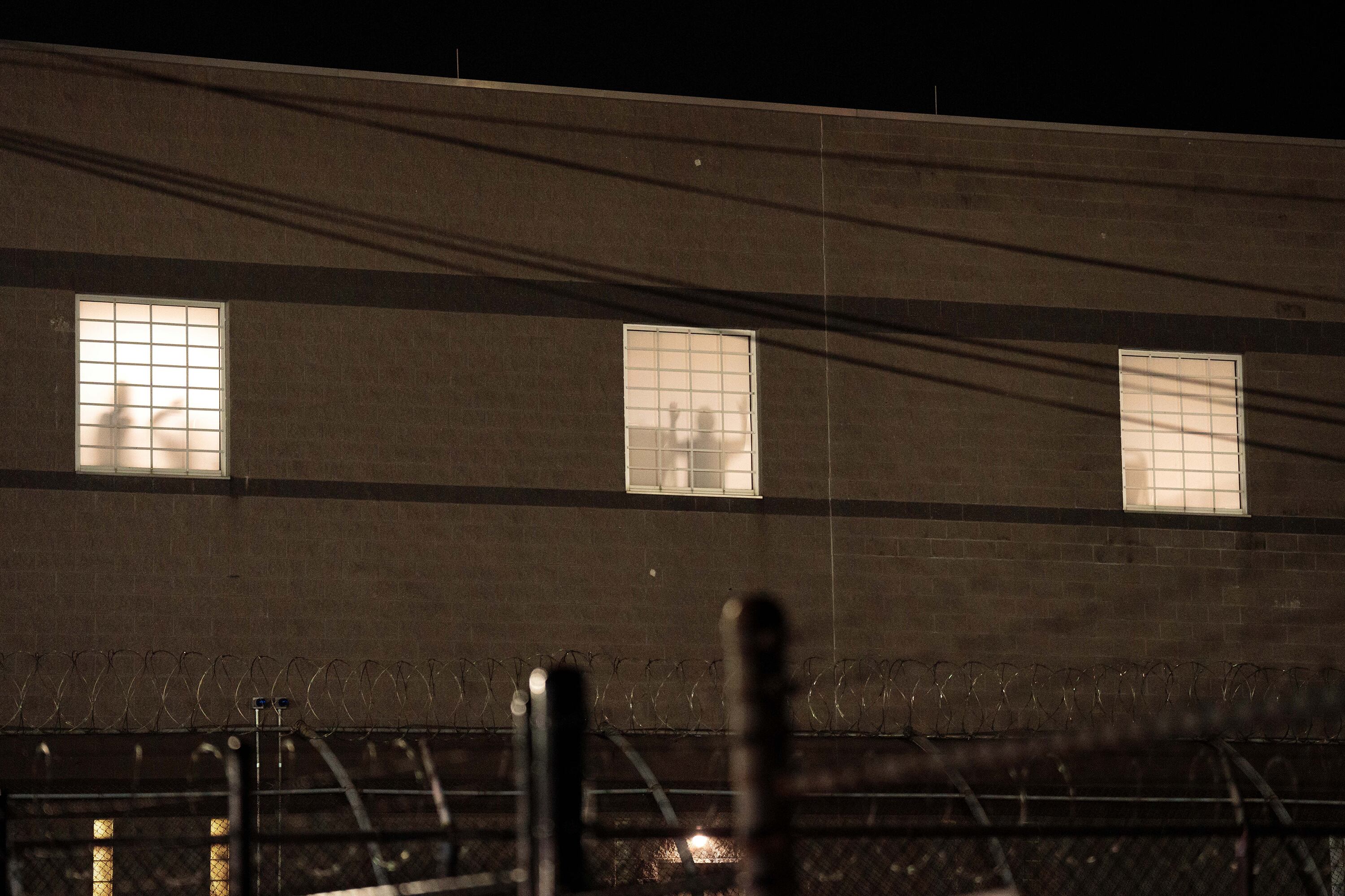A photograph of the outside of a large detention center with silhouettes of people detained standing in the windows.