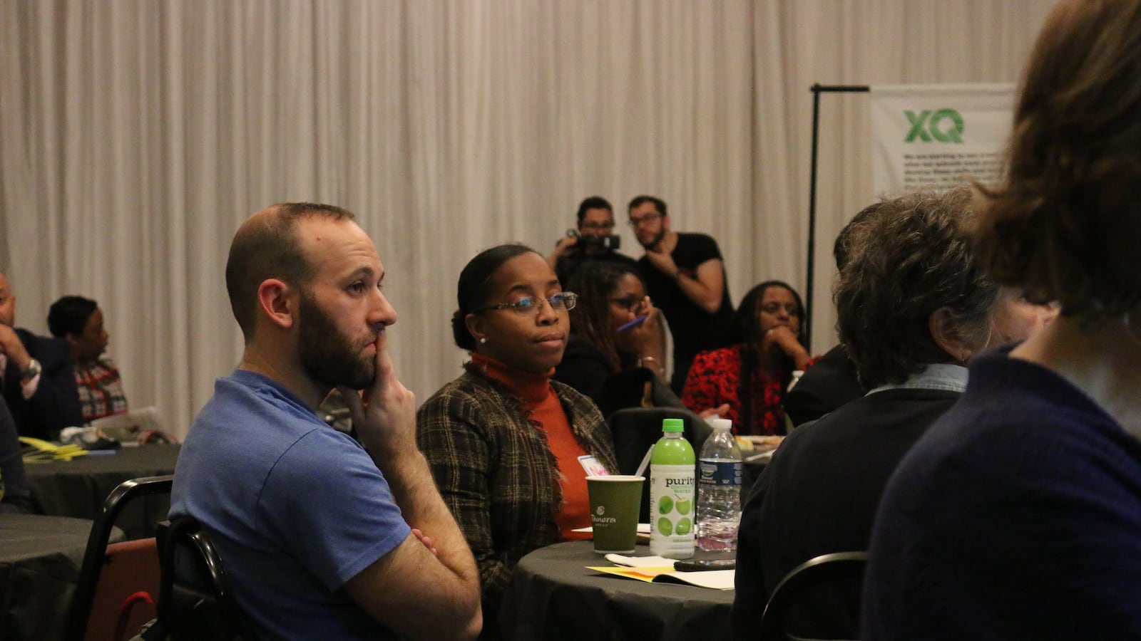 People sit around a table indoors, with a man wearing blue in the foreground.