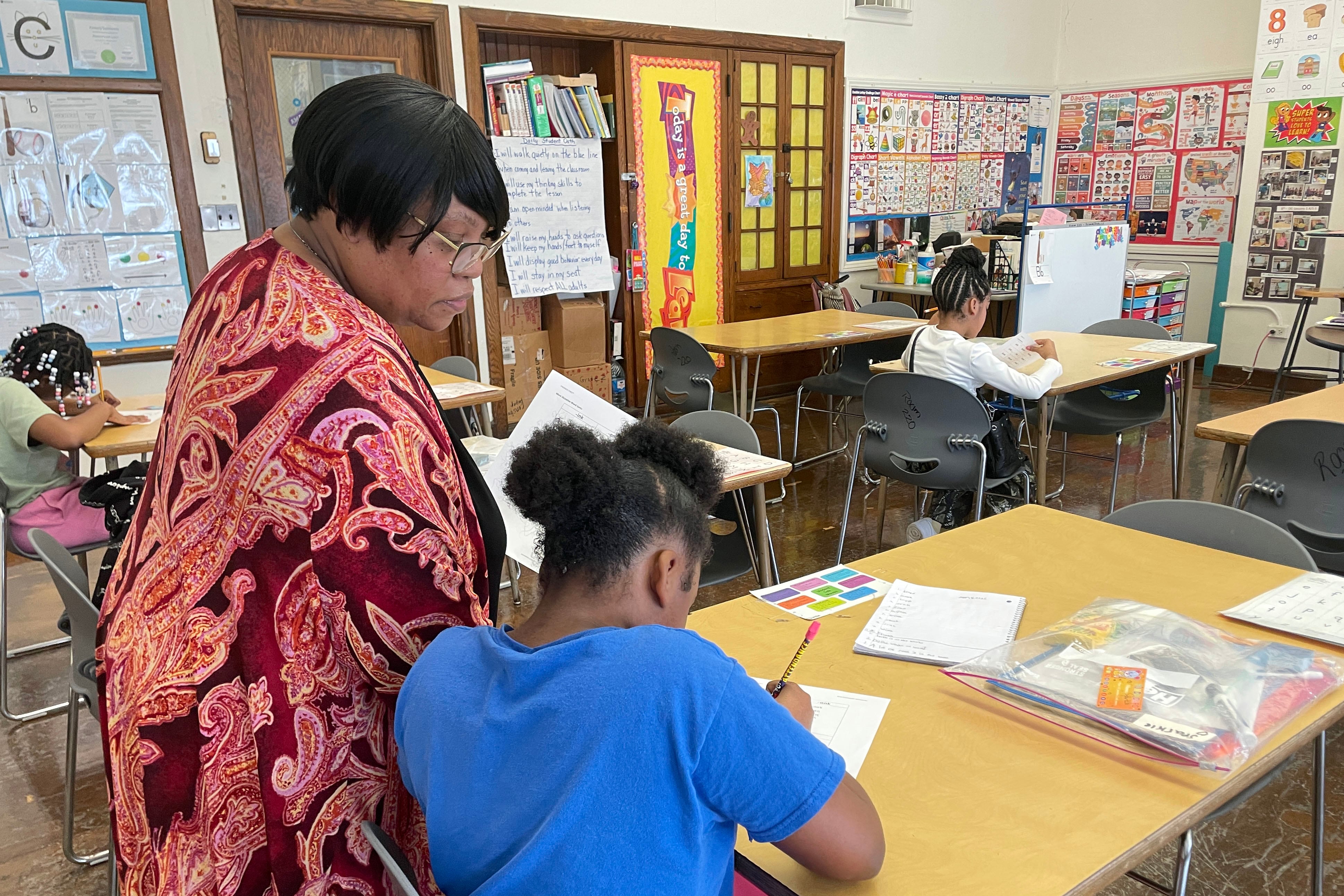 A Black woman helps a young student sitting at a desk in a classroom.