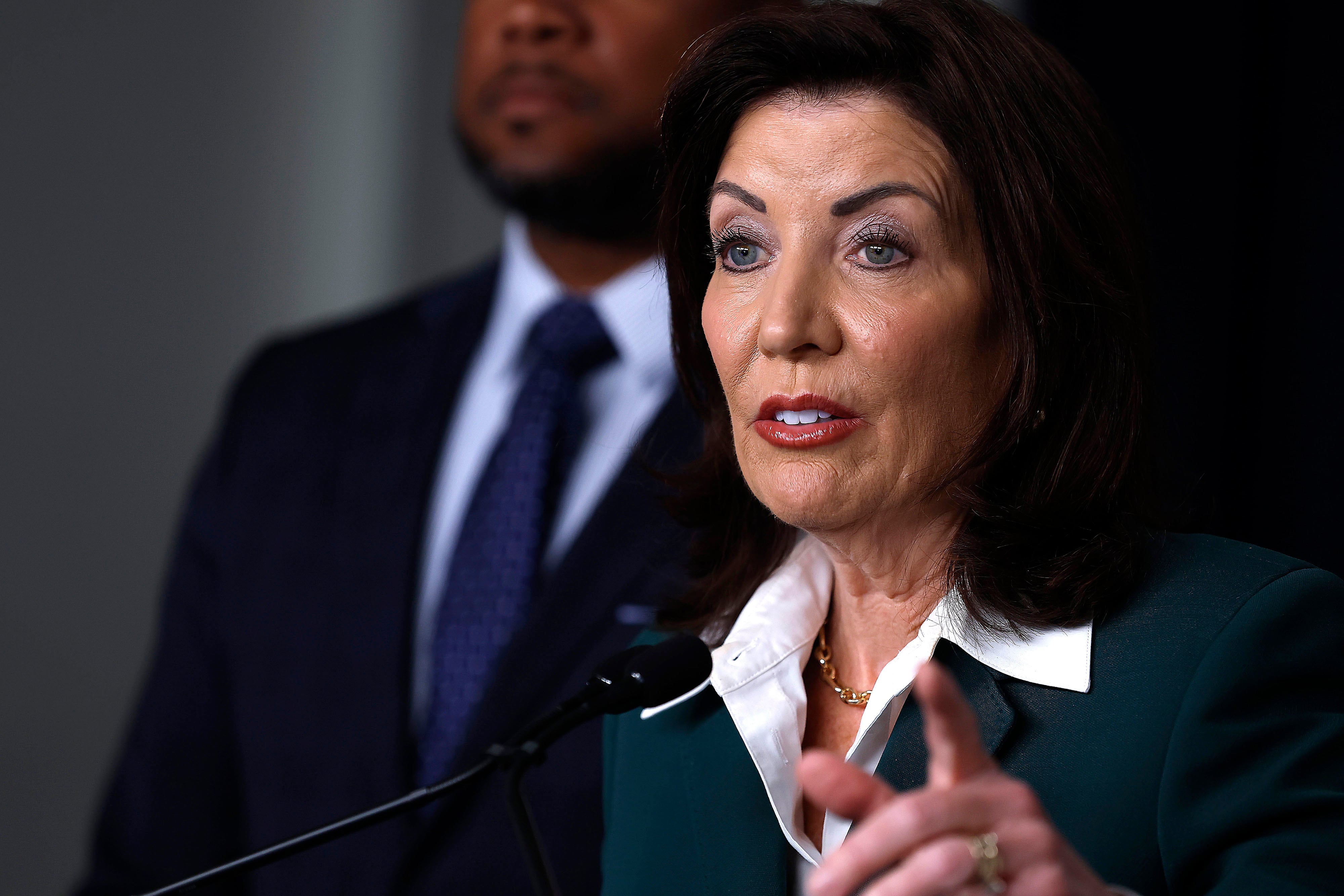A white woman in a suit speaks from a small microphone with a man in a suit standing behind her.