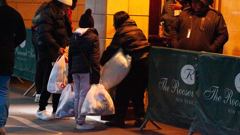Families stand in front of a hotel with their belongings in plastic bags.