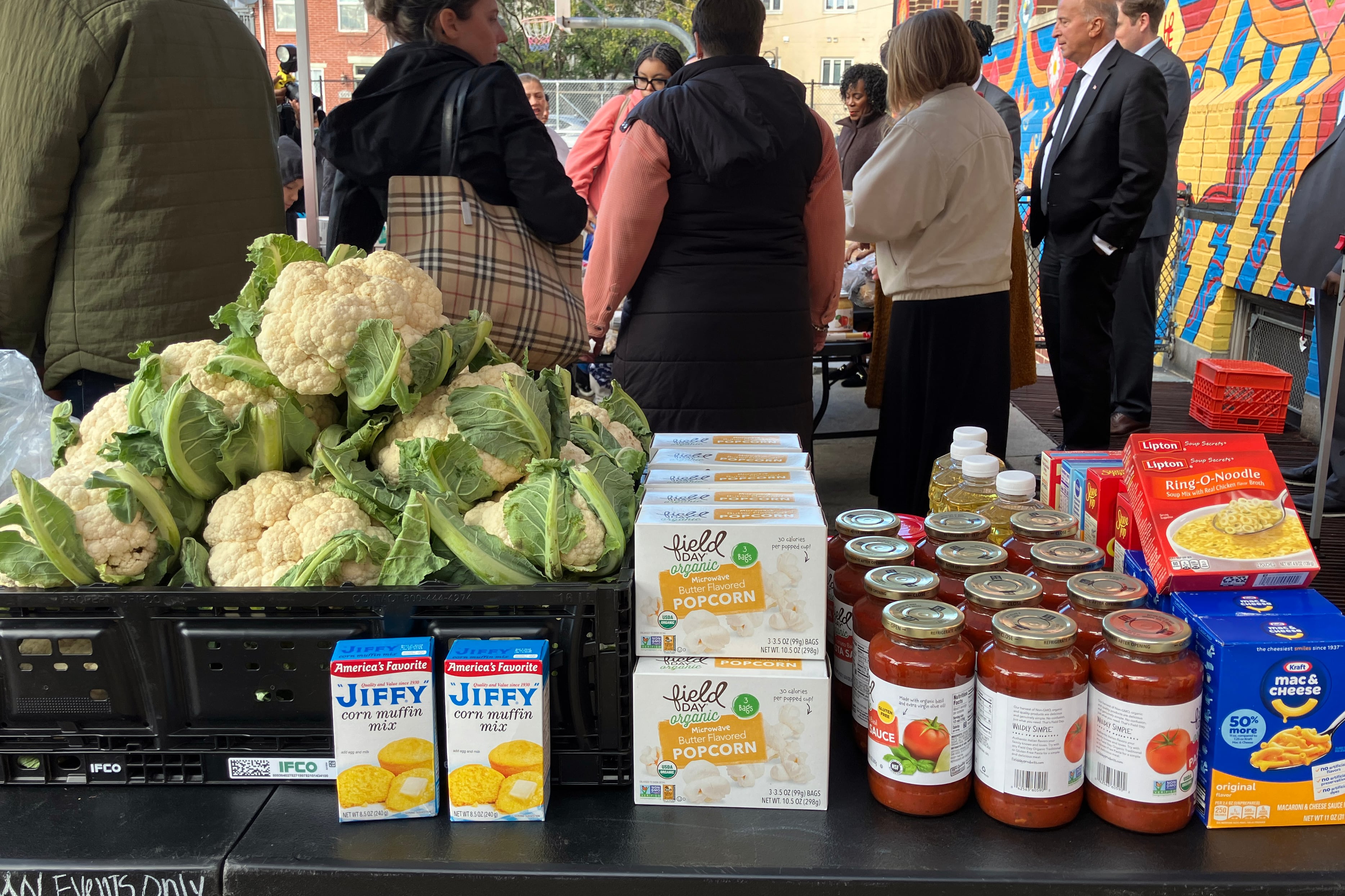 A photograph of a table full of shelf stable goods and cauliflower with a group of people in the background.