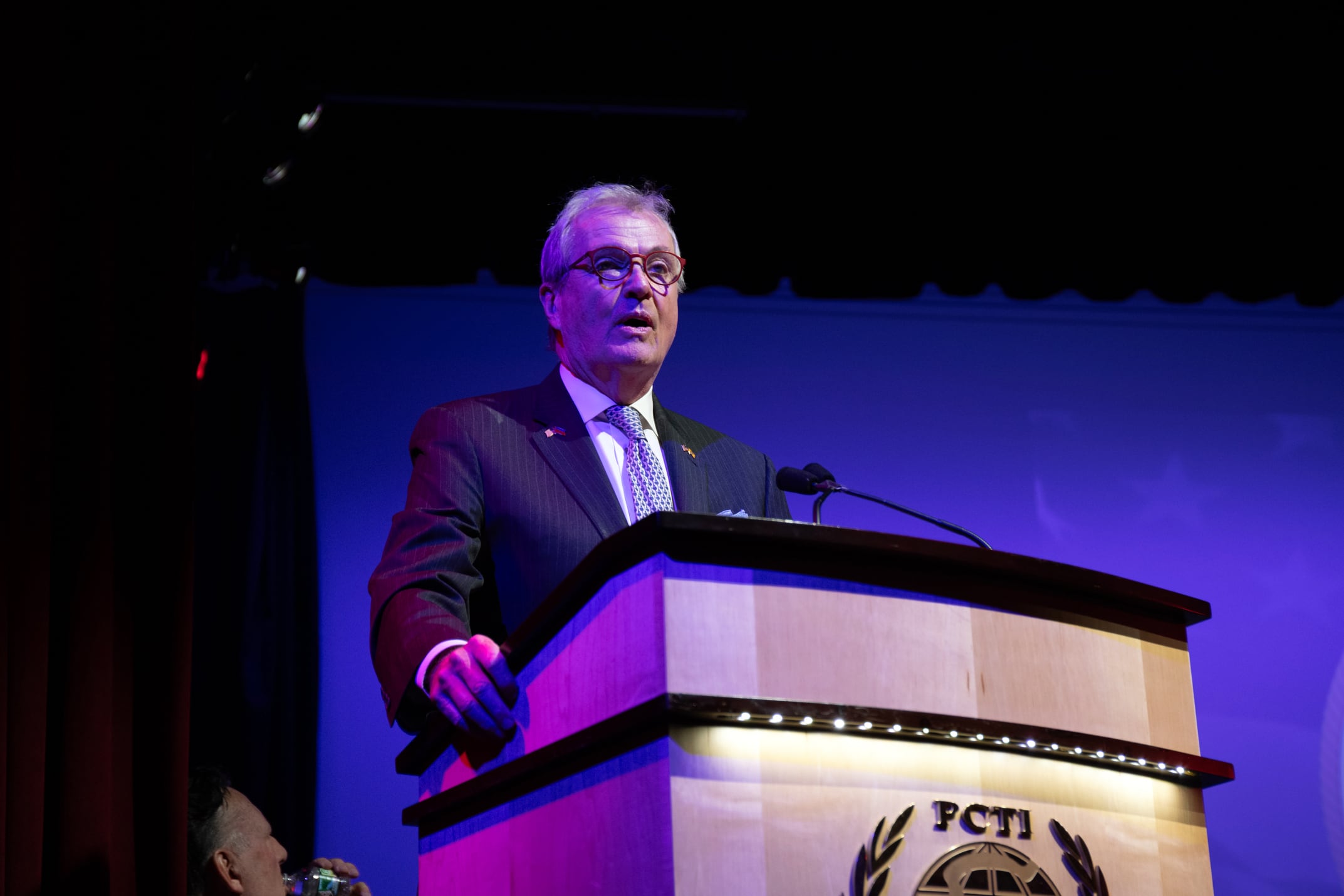 A man in a suit speaks from behind a podium and in front of a blue backdrop.