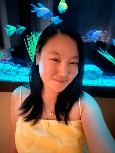 A high school student with short black hair and wearing a yellow top stands in front of a fish tank.