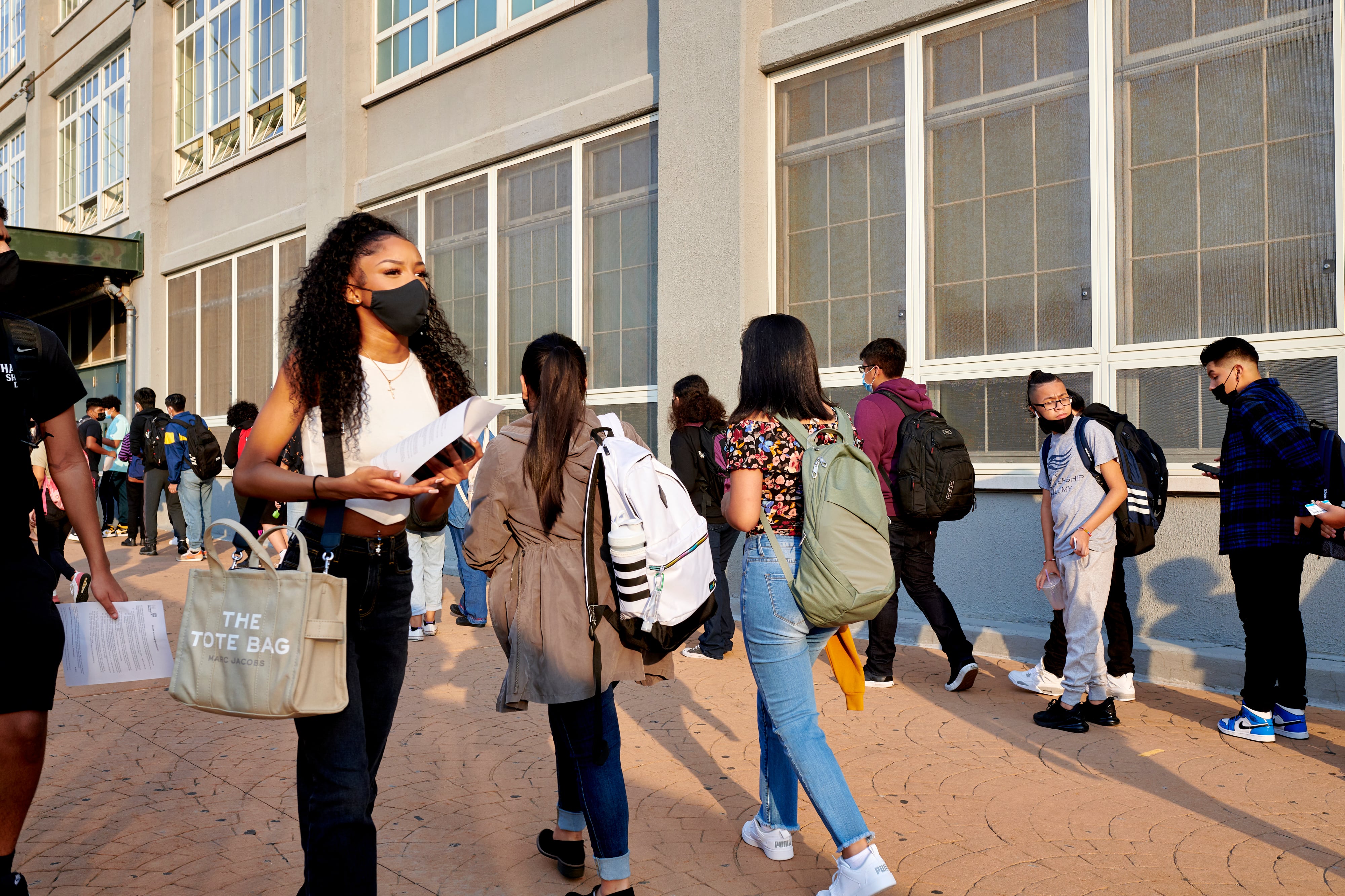 A teenage girl with long curly black hear wearing a black face mask, white sleeveless shirt and black pants and a back pack walks away from a school building as other teens line up to walk inside.