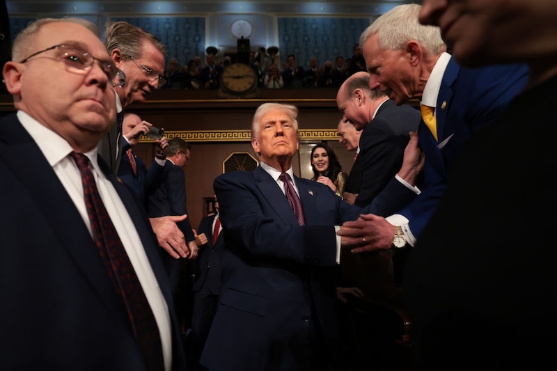 U.S. President Donald Trump, an older man in a dark suit and red tie, walks through a crowd of other men in suits.