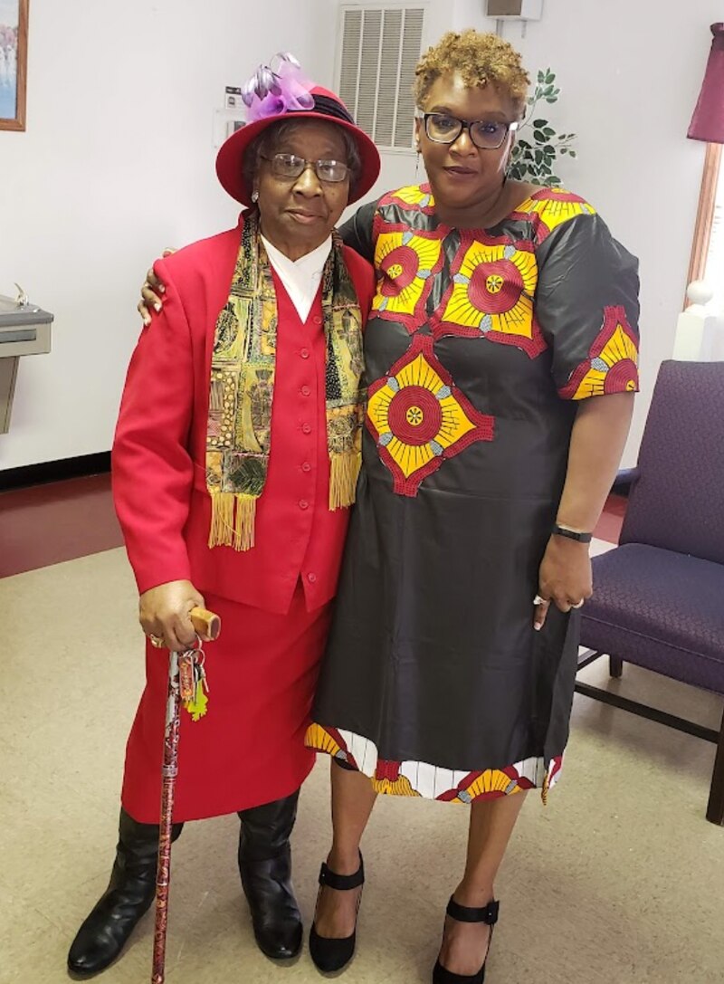 Two women pose for a photo. The woman on the left wears a red suit and hat. The woman on the right wears a black, red, and yellow dress.