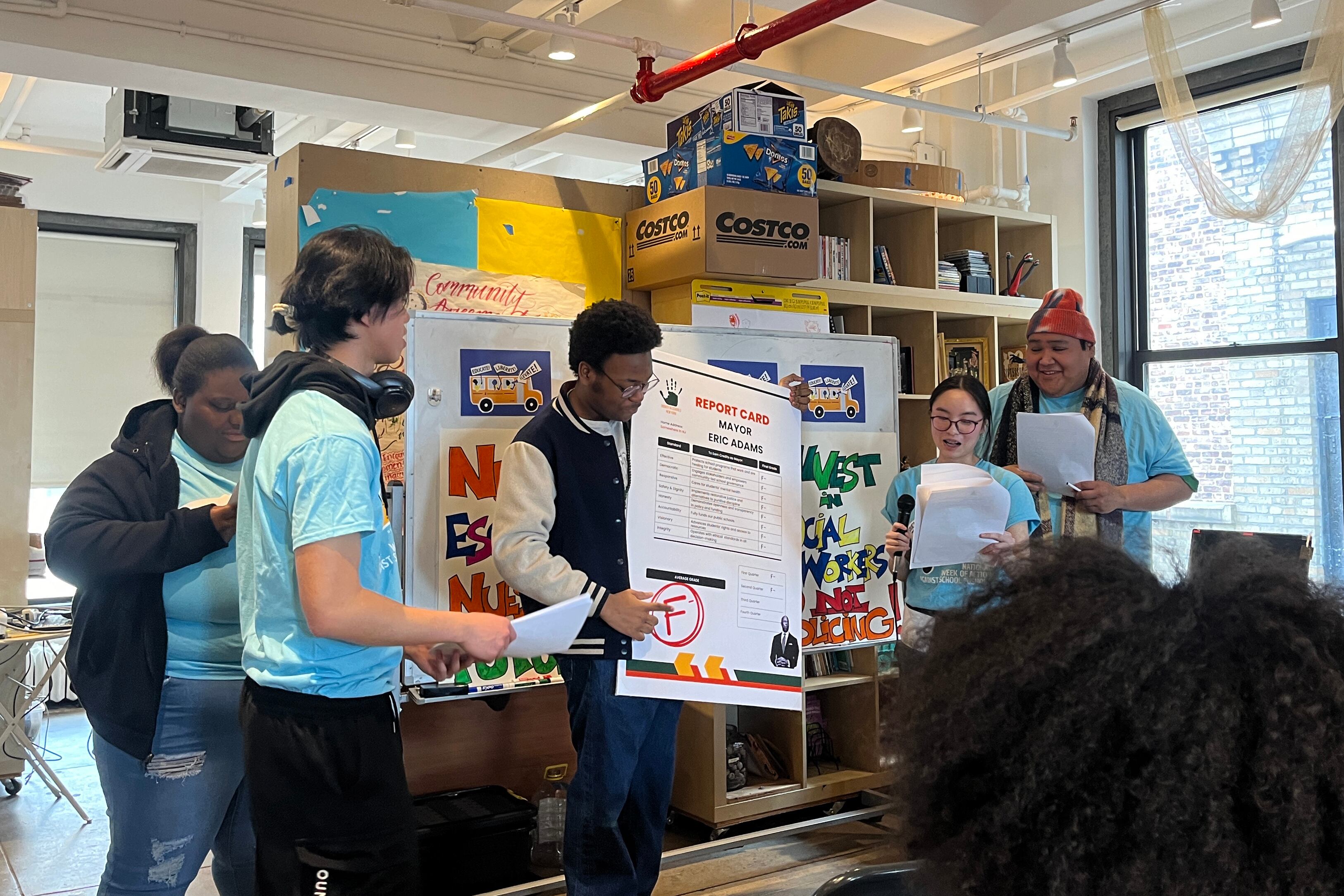 Five high school students all but one wearing light blue t-shirts stand in front of a crowd (not pictured) holding a poster with information on it.