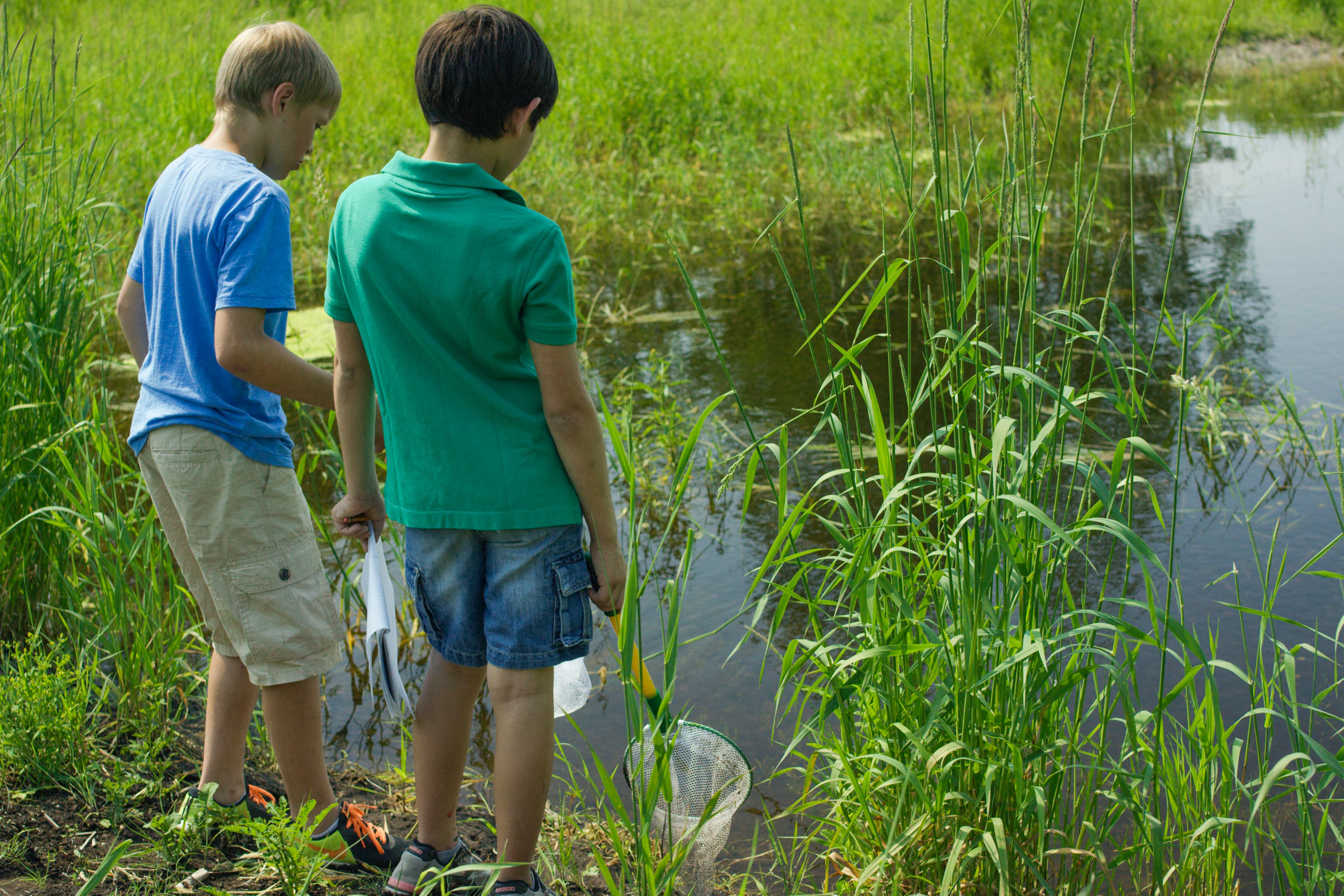 Two boys in shorts stand near a pond.