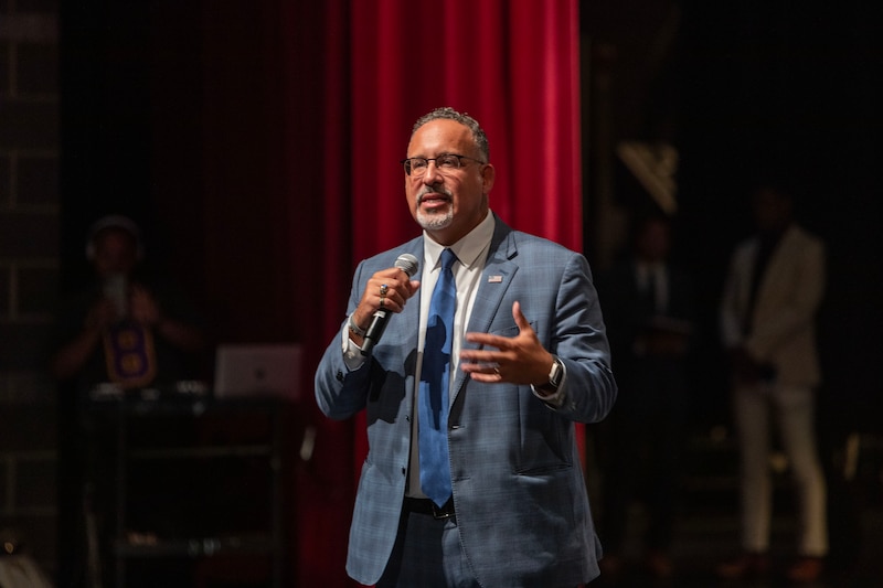 A man wearing a blue suit stands on stage and speaks into a microphone.