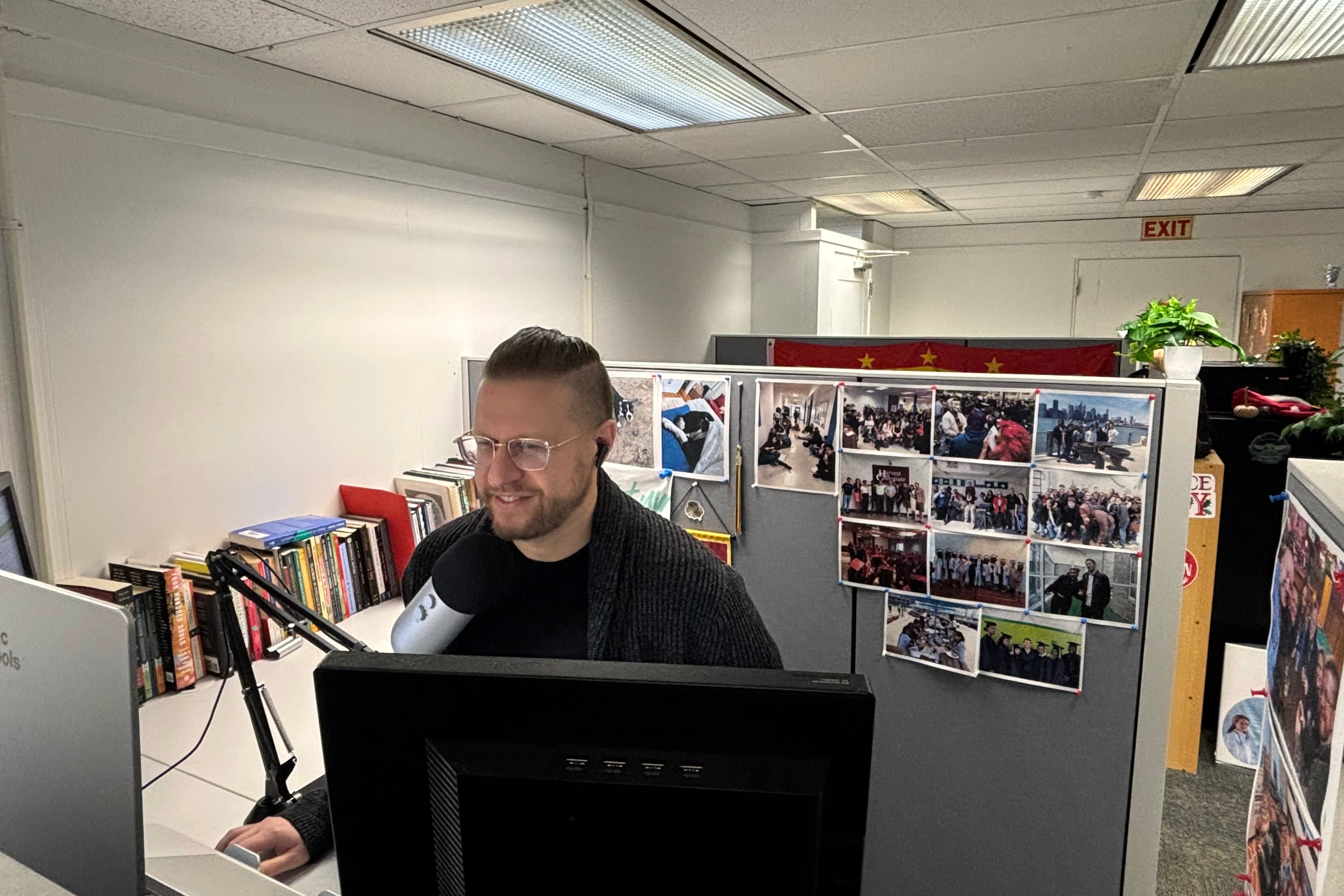 A white man works from two computers in a cubicle with photos lining two sides of his work space.