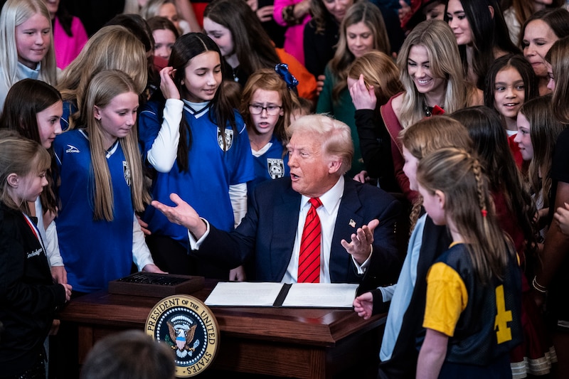 A man in a suit sitting at a wooden desk while a large group of young girl athletes stand around.