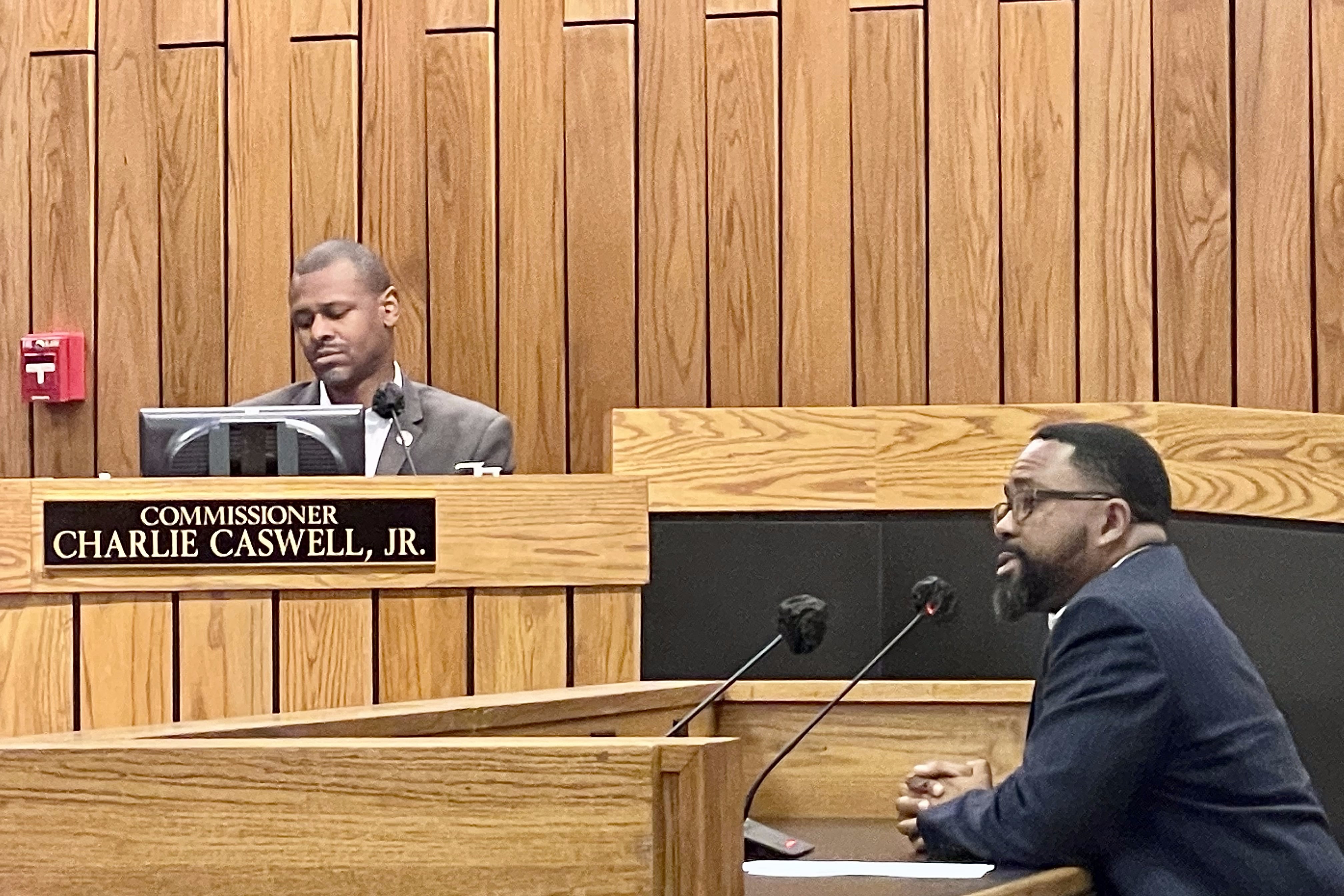 A man sitting at a wooden desk speaks into a microphone